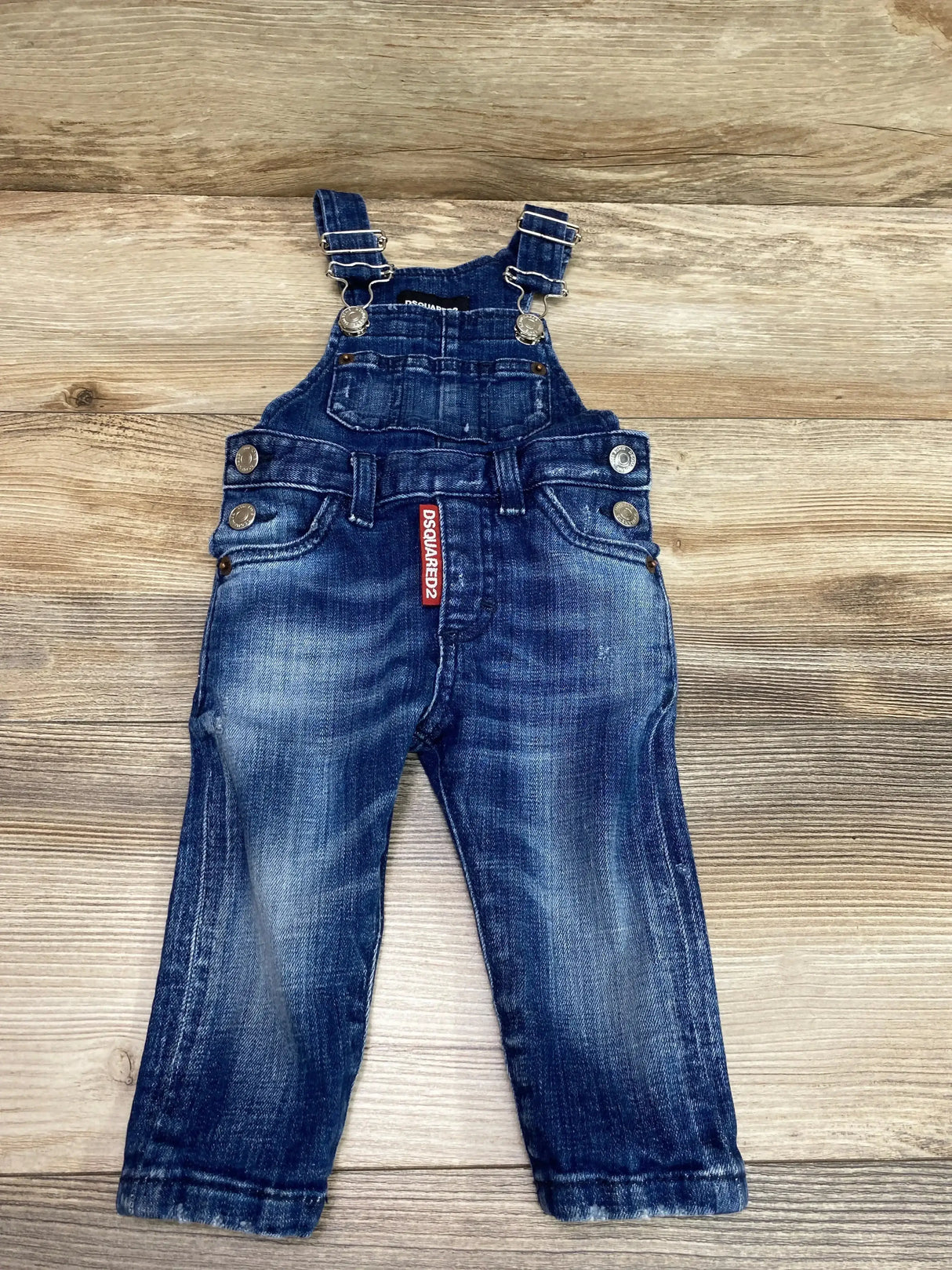 Front flat-lay of Dsquared2 baby denim jumpsuit 9M with adjustable buckle straps, faded wash and red DSQUARED2 tag on wood