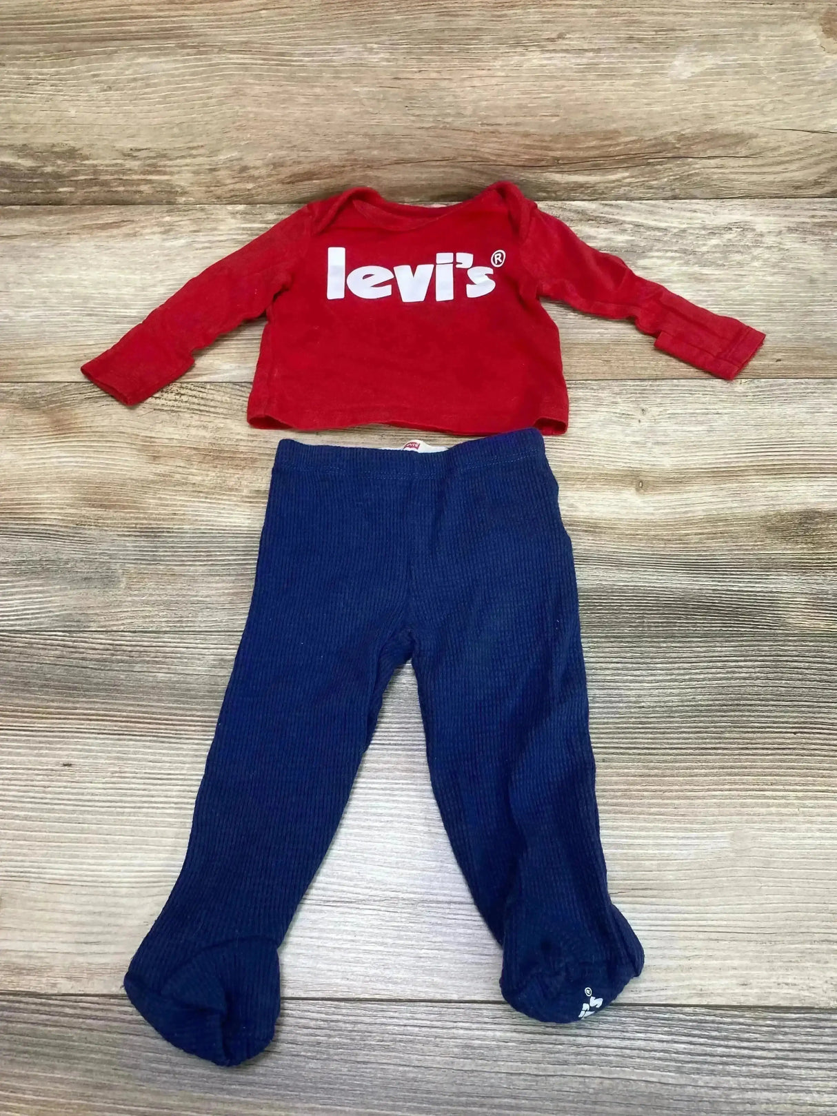Flat lay of Levi’s red long-sleeve baby shirt with white logo and navy footed pants on wood floor, size 3 months