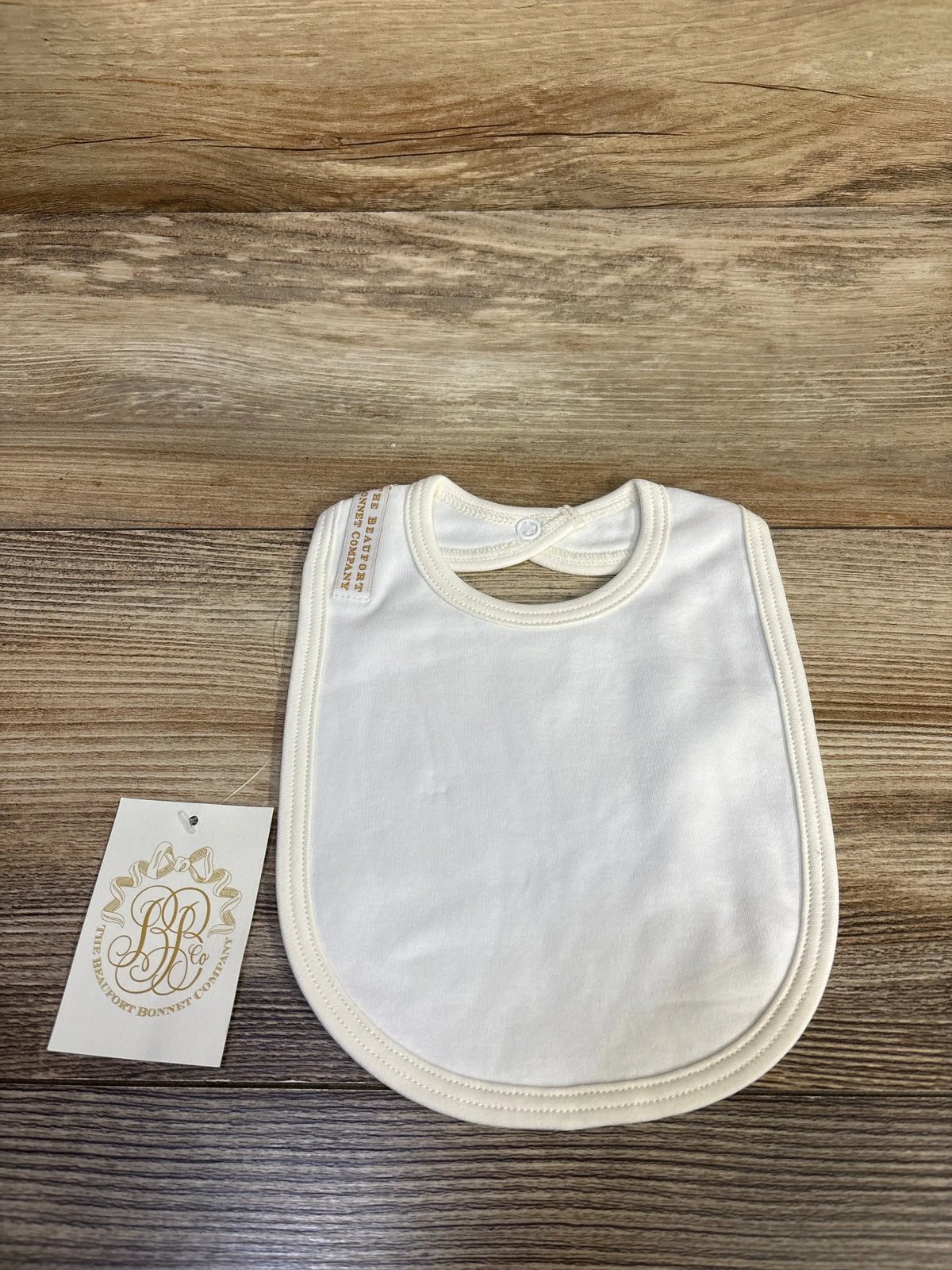 Front view of The Beaufort Bonnet Company Burp Me Bib showcasing white fabric and neck snap on wood-grain background