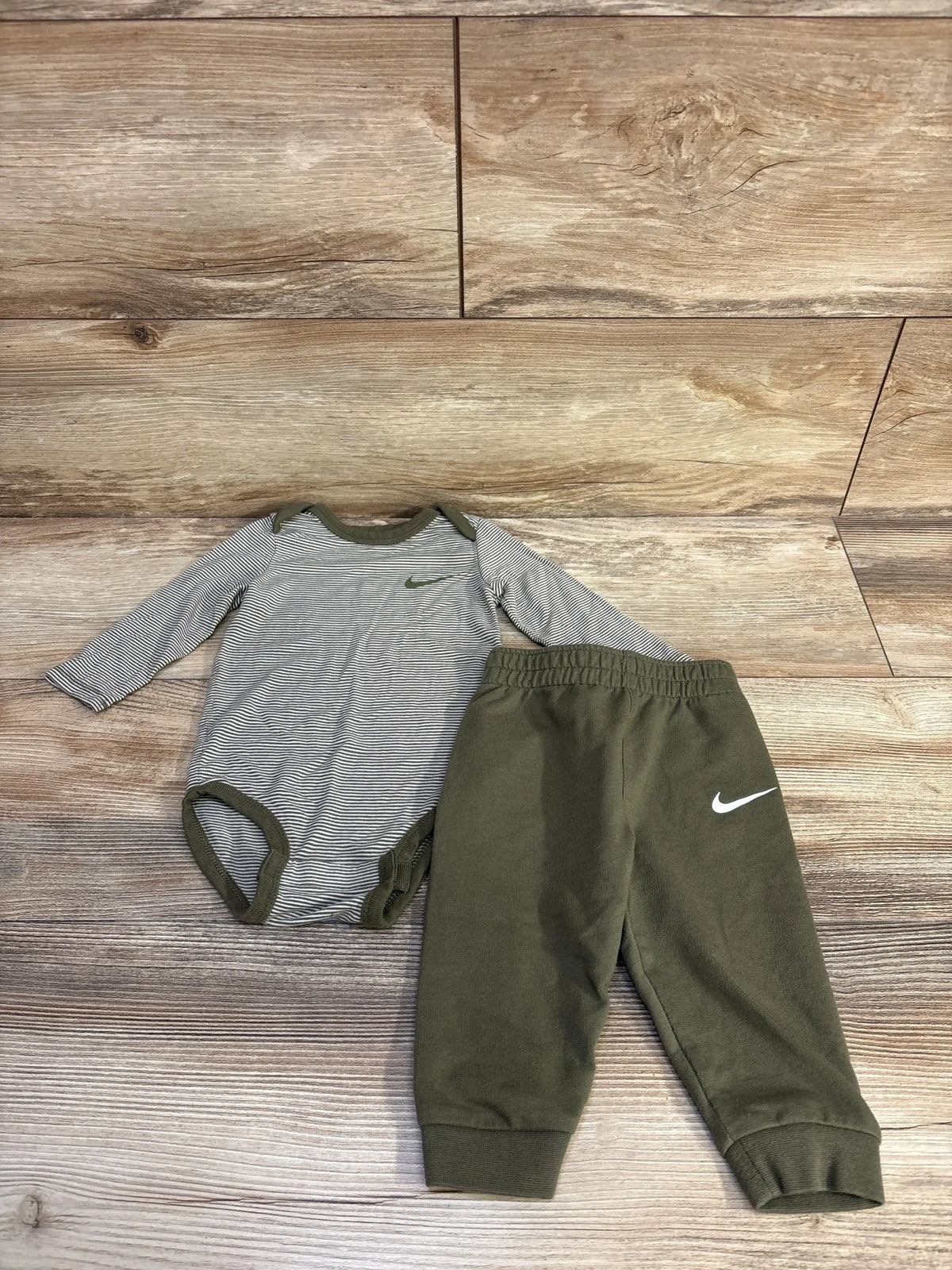 Front view of Nike 2-piece bodysuit and bottoms set, size 6 months, showcasing Swoosh logos on a wood-look background