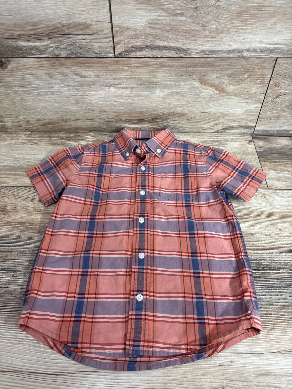 Top-down view of size 5T orange plaid Janie and Jack short-sleeve poplin button-down shirt showcasing button front, chest pocket, and curved hem on wood background