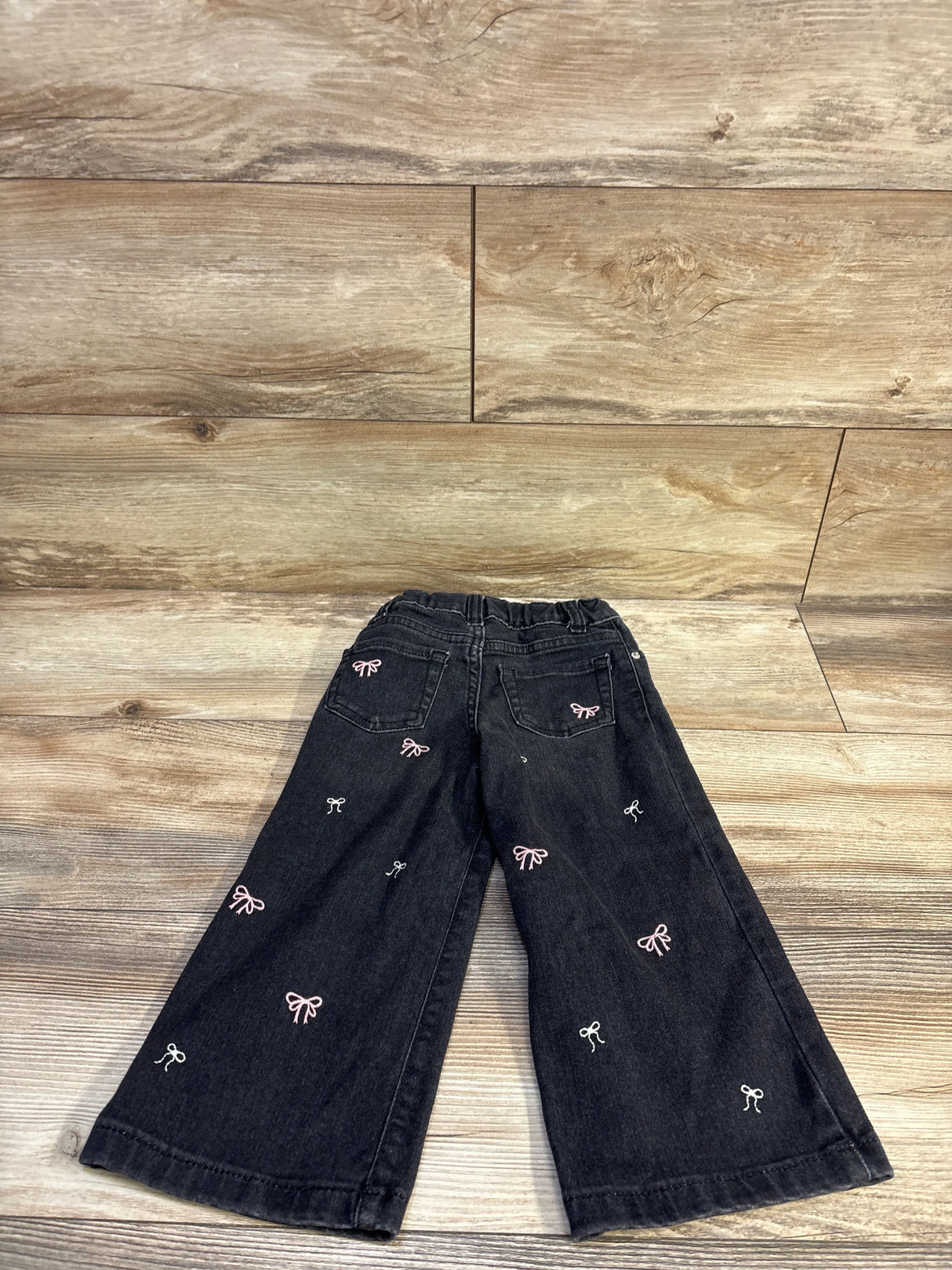 Back of Cat and Jack wide-leg black toddler jeans showing pink and white embroidered bows on wood floor, size 3T