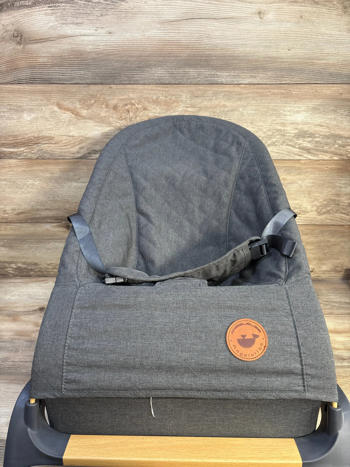 Front view of ANGELBLISS portable baby bouncer showcasing quilted grey seat, safety harness, and wooden front bar on wood plank background