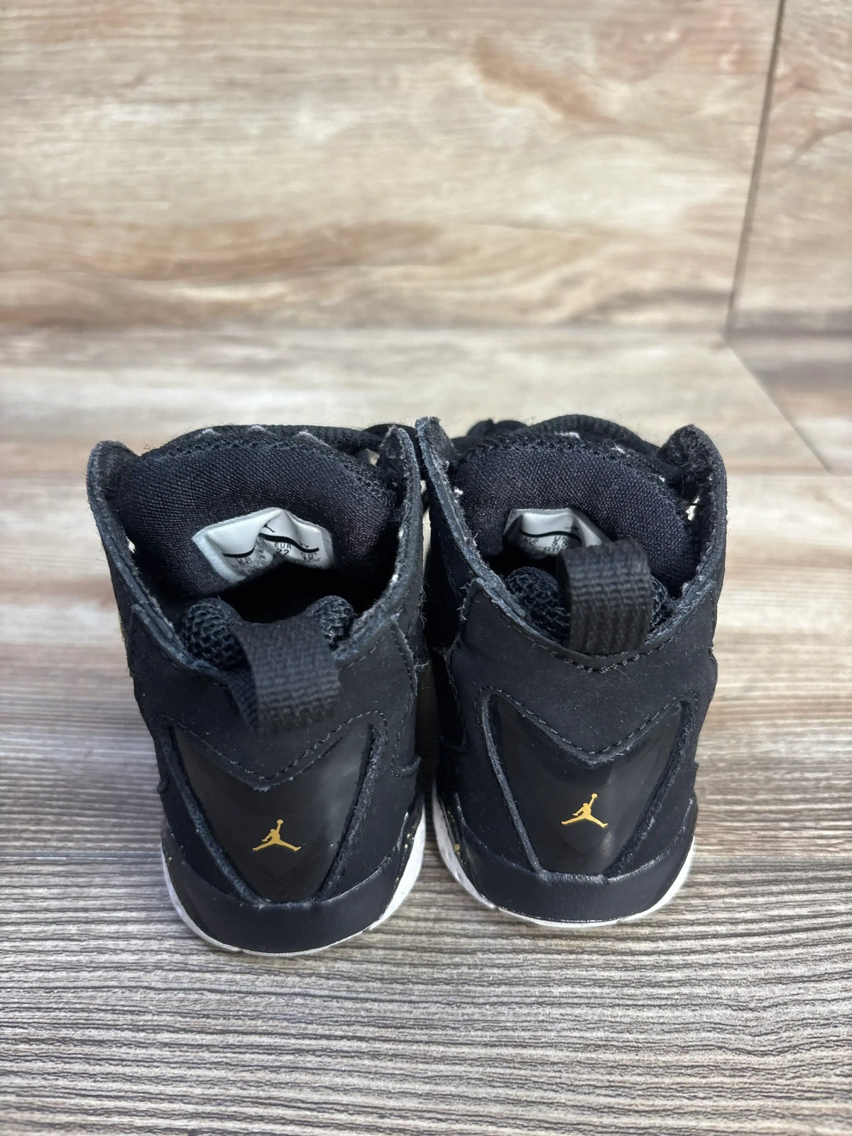 Rear view of Air Jordan True Flight BT black sneakers showcasing gold Jumpman logos and white rubber outsoles on a wood-look floor