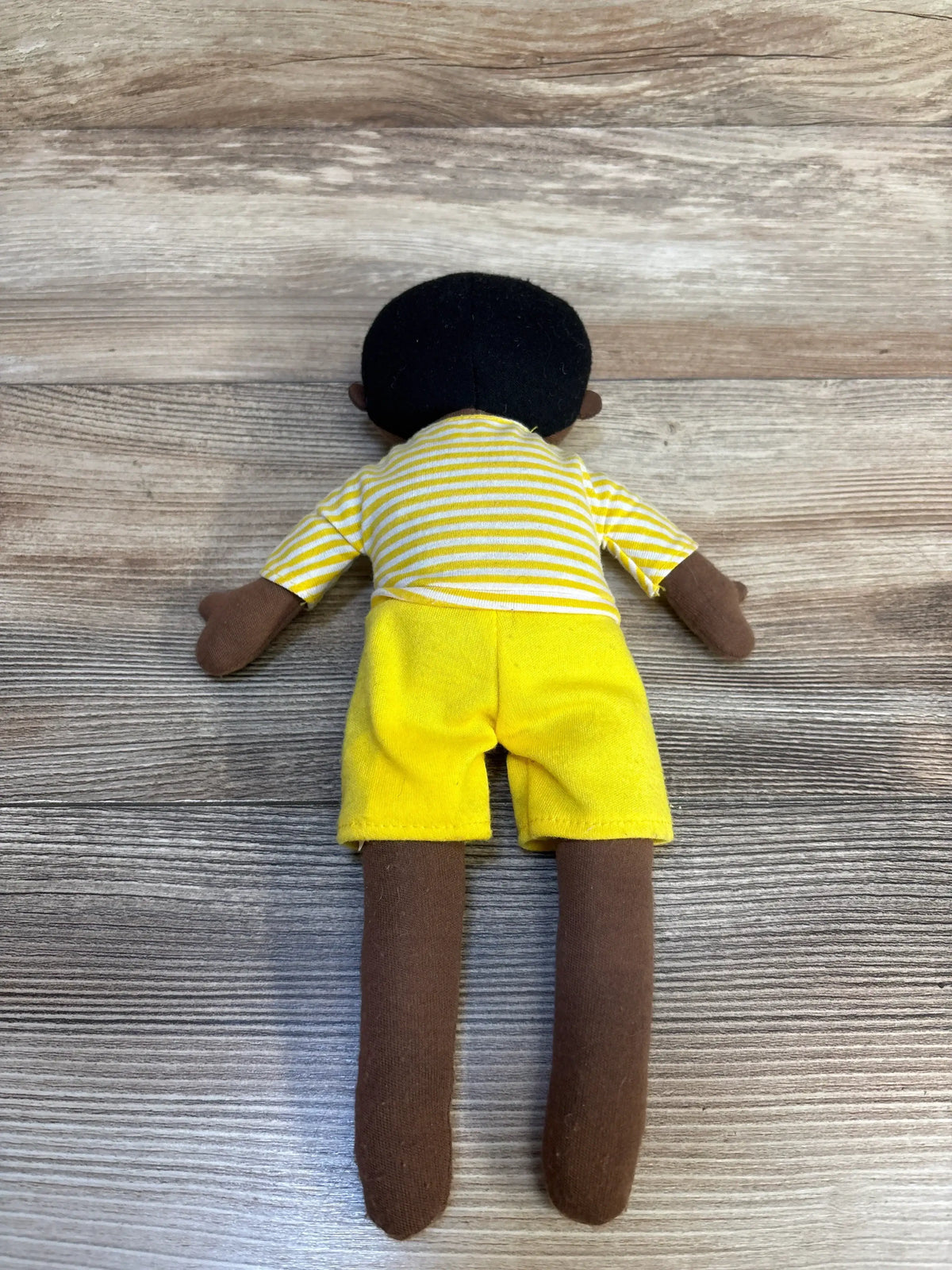 Back view of Cloud Island Plush Doll showing brown fabric skin, black felt hair, yellow-striped shirt and bright yellow shorts on wood surface