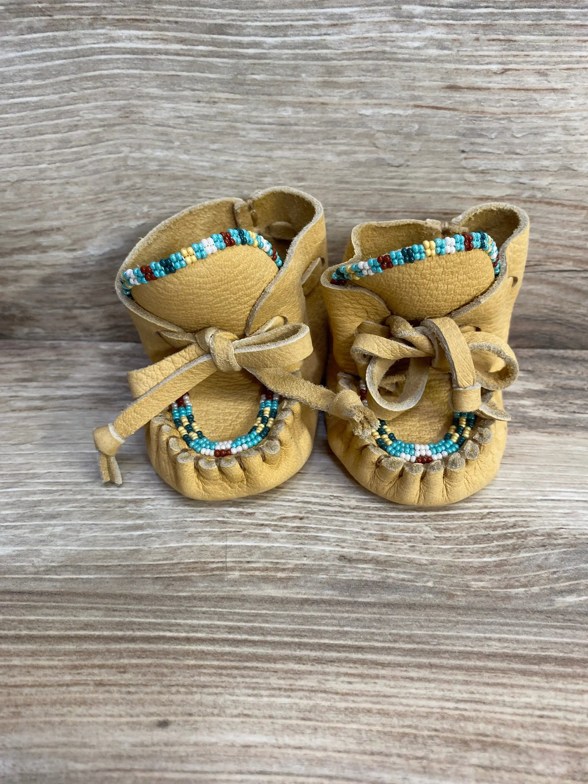 Front view of Baby Bootie Beaded Leather Moccasin Slippers sz 2c showcasing tan leather with turquoise red and white beaded trim and suede ties with wood plank background