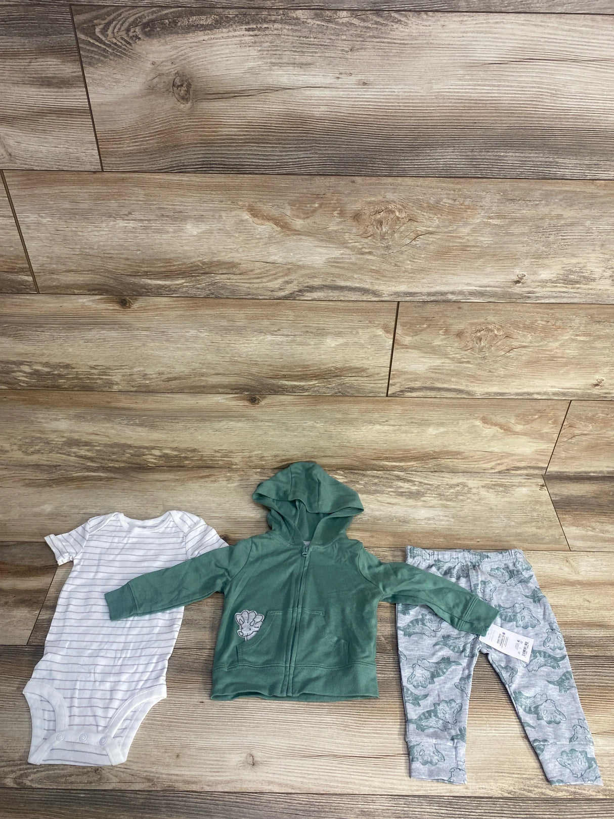 Infant clothing set displayed on wood floor including white striped bodysuit, green zip-up hoodie, and gray dinosaur print leggings