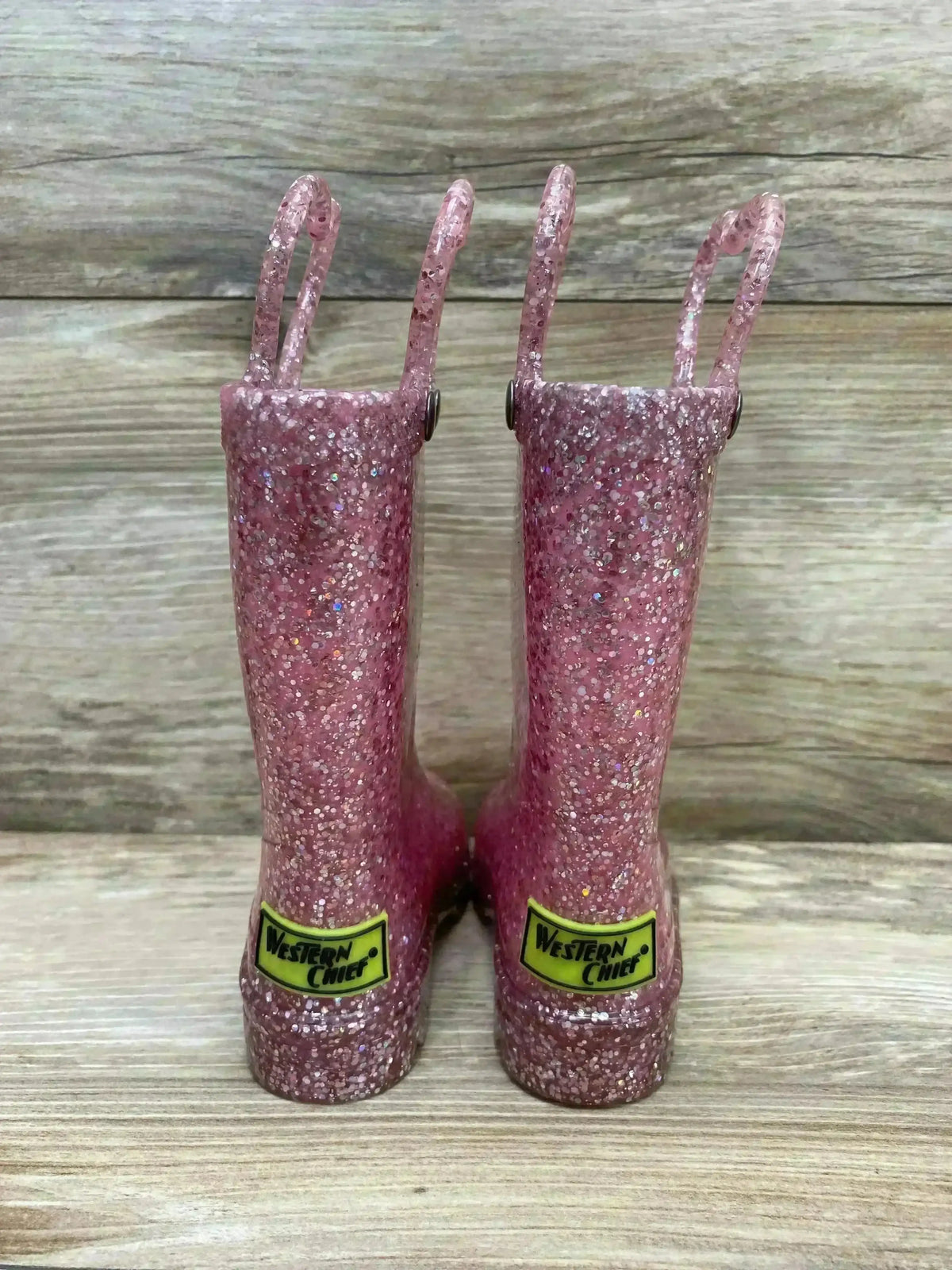 Back view of Western Chief pink glitter light up rain boots showcasing pull tabs and logo patch on wood plank floor sz 5c