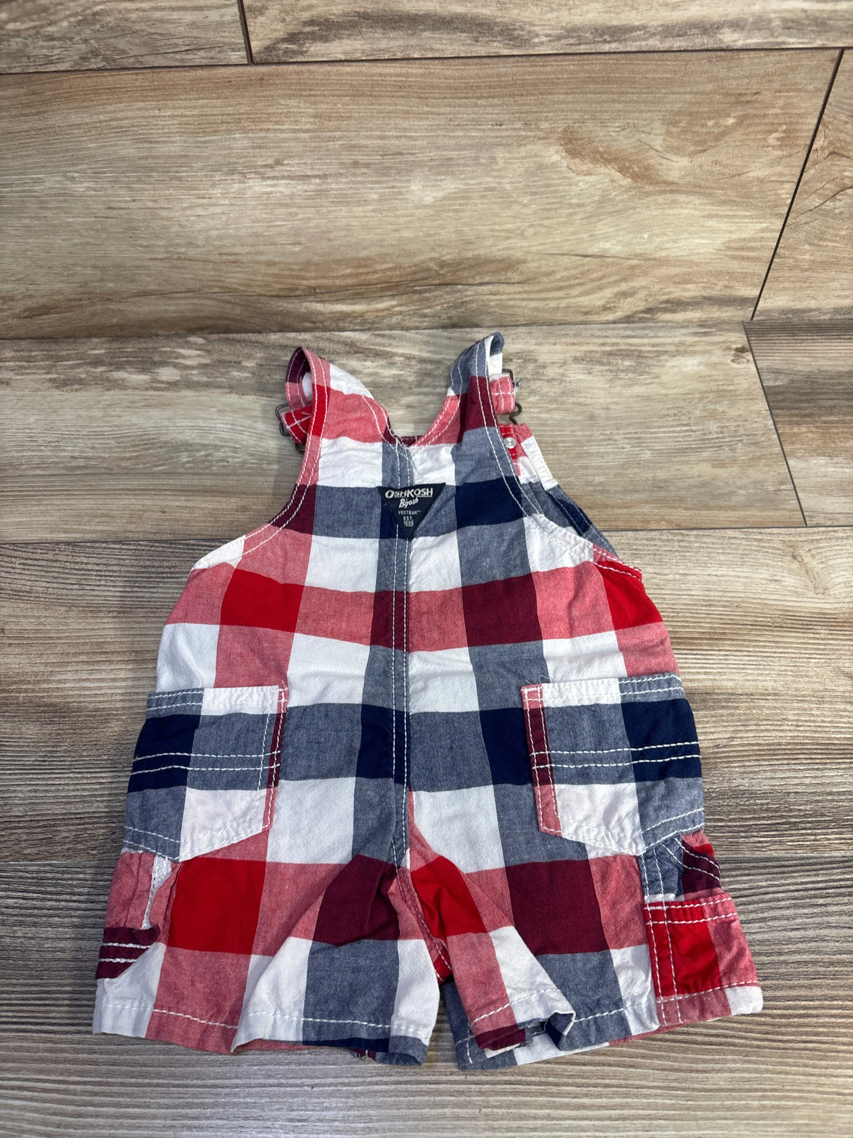 Front view of OshKosh shortalls size 12m showcasing red white and blue plaid pattern with adjustable straps and front pocket on wood plank floor