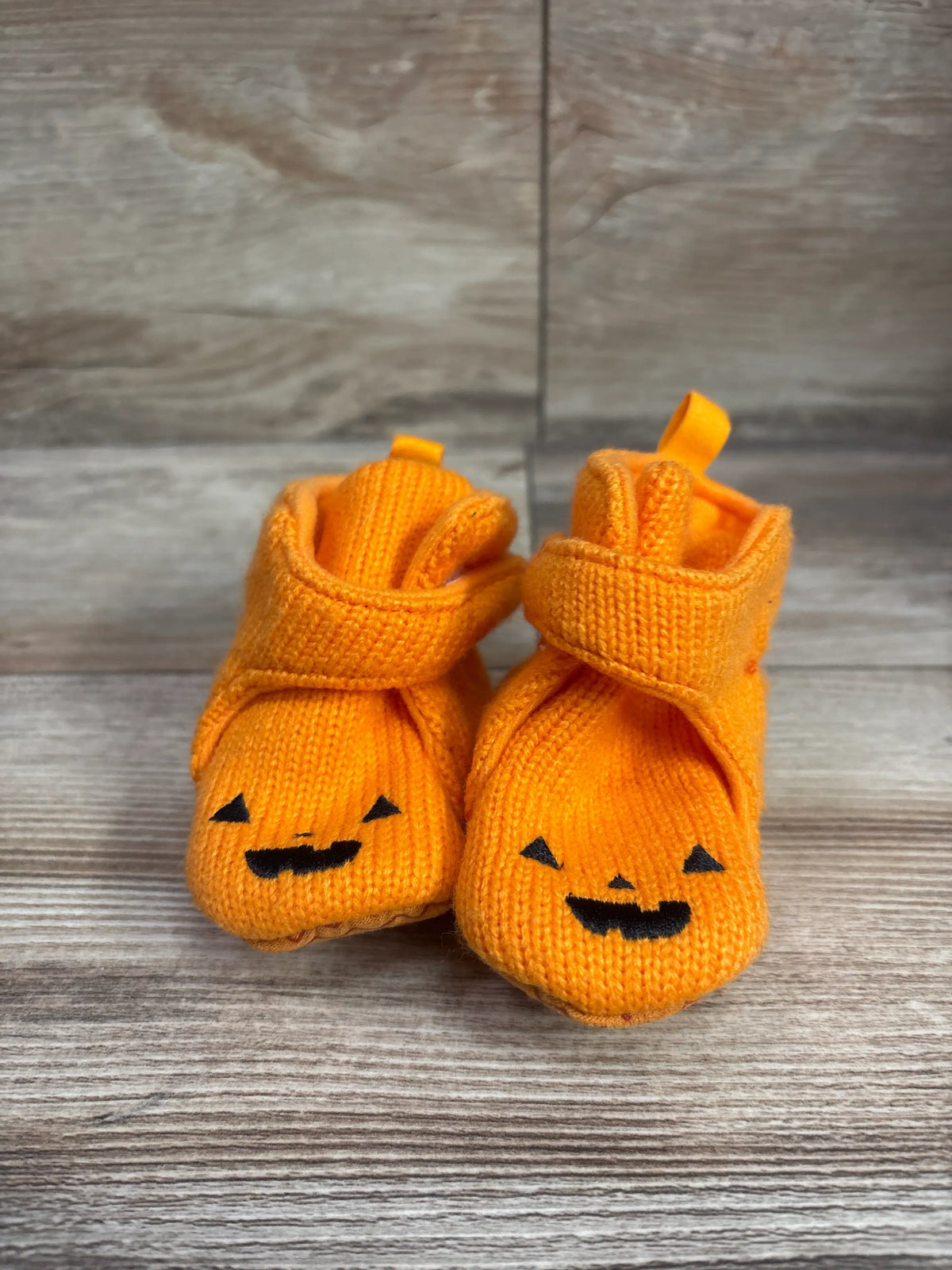 Front view of Cat & Jack pumpkin crib booties in orange, showcasing jack-o-lantern faces and foldover cuffs on a wood-grain background