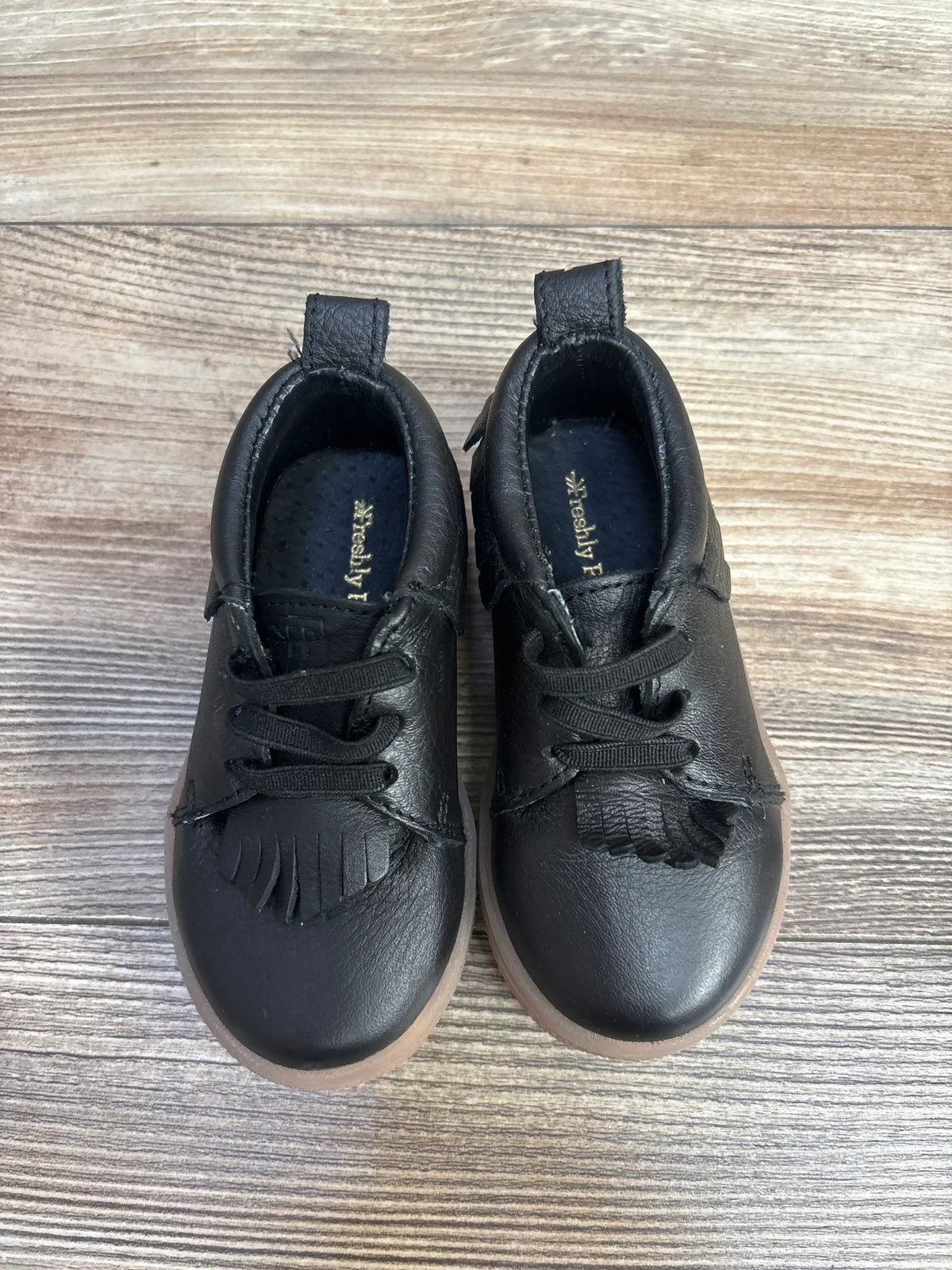Top-down of Freshly Picked slip-on mid sneakers (black, size 5C) showing fringed tongue and brown sole on wood floor