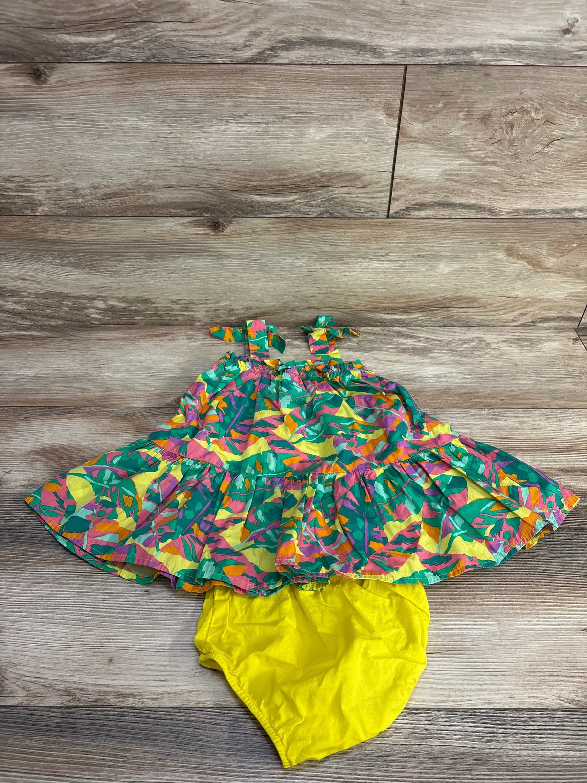 Top-down view of sleeveless multicolor tiered baby dress with shoulder tie straps and matching yellow bloomers on wood floor