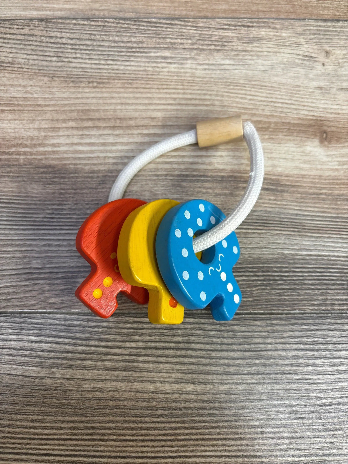 Angled view of Plan Toys Baby Key Rattle showcasing three colorful wooden keys on a white cord with a woodgrain background