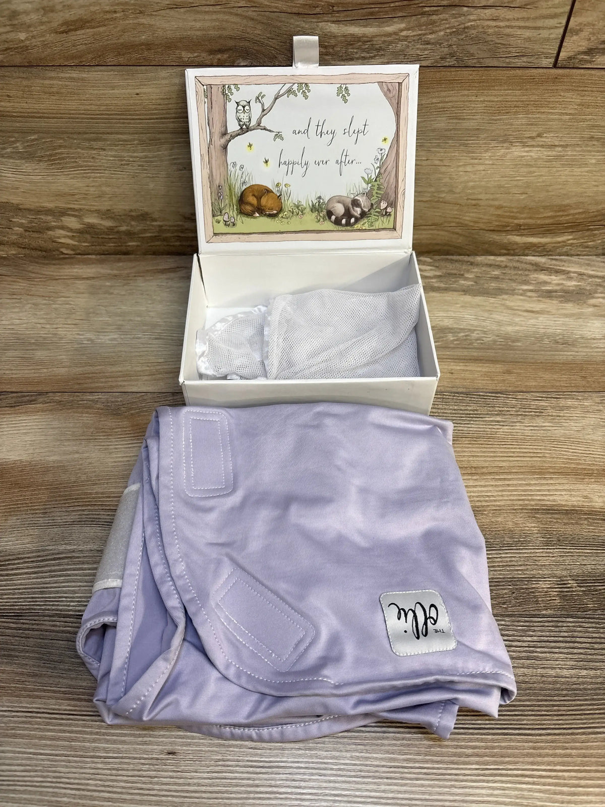 Front view of The Ollie World lavender swaddle showcasing custom closure tabs with an open woodland-themed gift box behind it