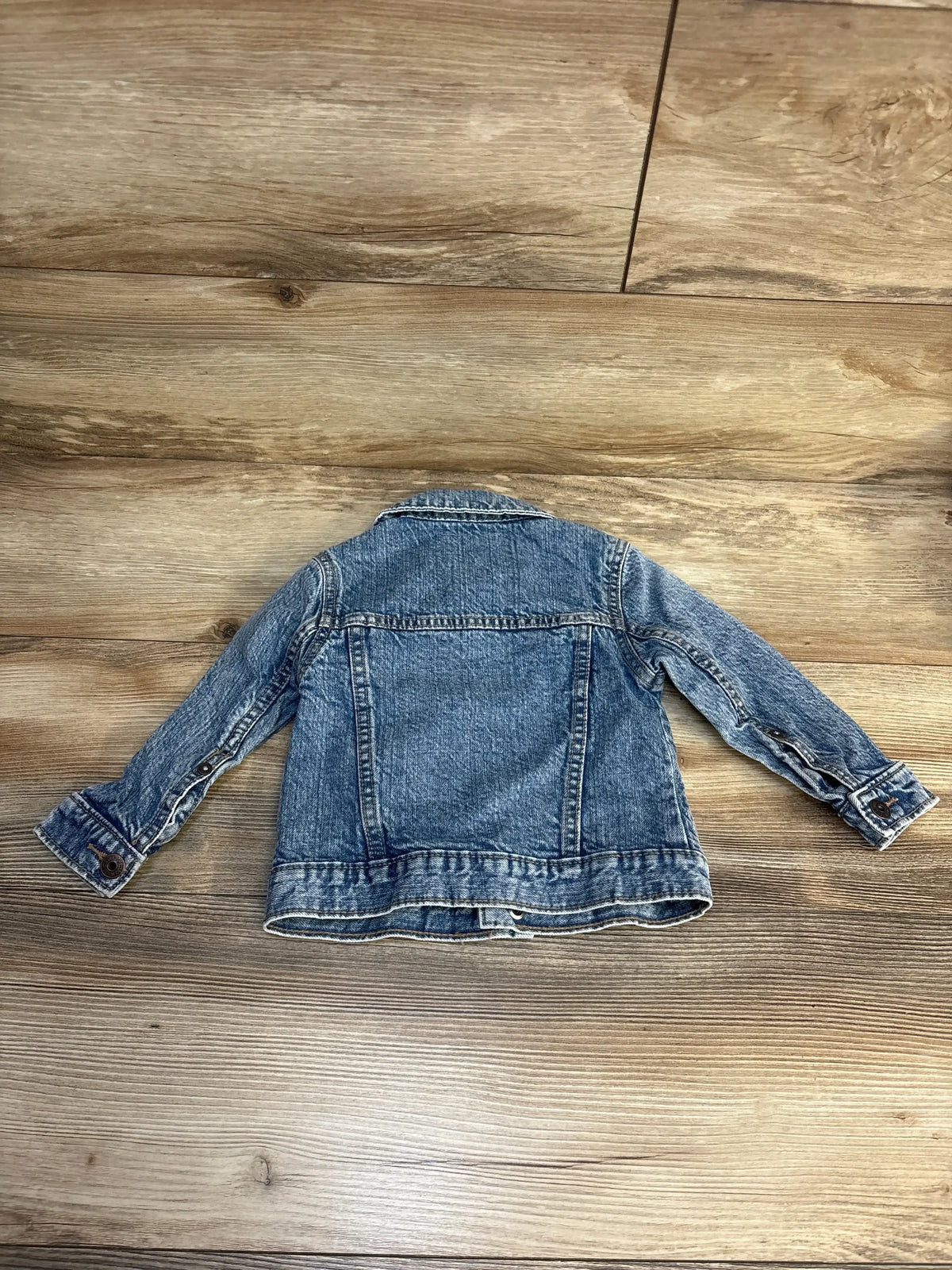 Back view of OshKosh light blue toddler denim jacket size 18 months showing button cuffs and seam details on wood floor