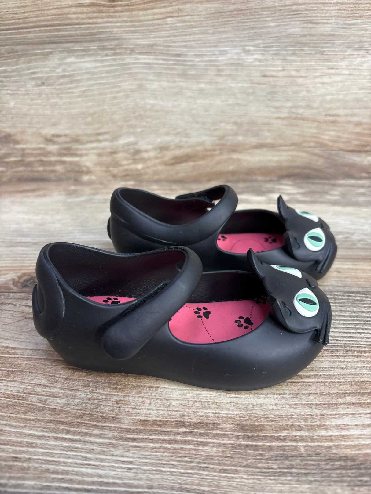 Side view of Mini Melissa Ultragirl II flats in black showing cat-face toe with green eyes and pink paw print insoles on wood background