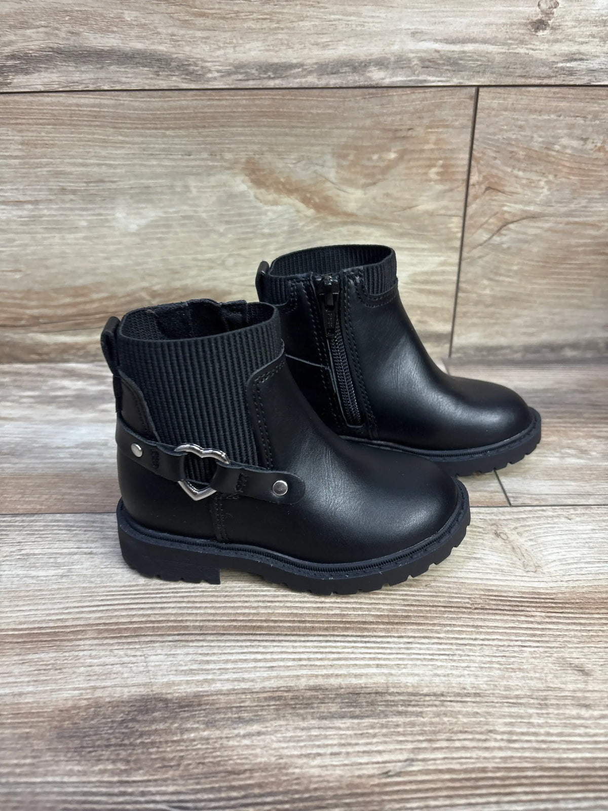 Side view of toddler black faux leather moto boots 6C showing side zipper, ribbed cuff, heart metal strap on wood floor