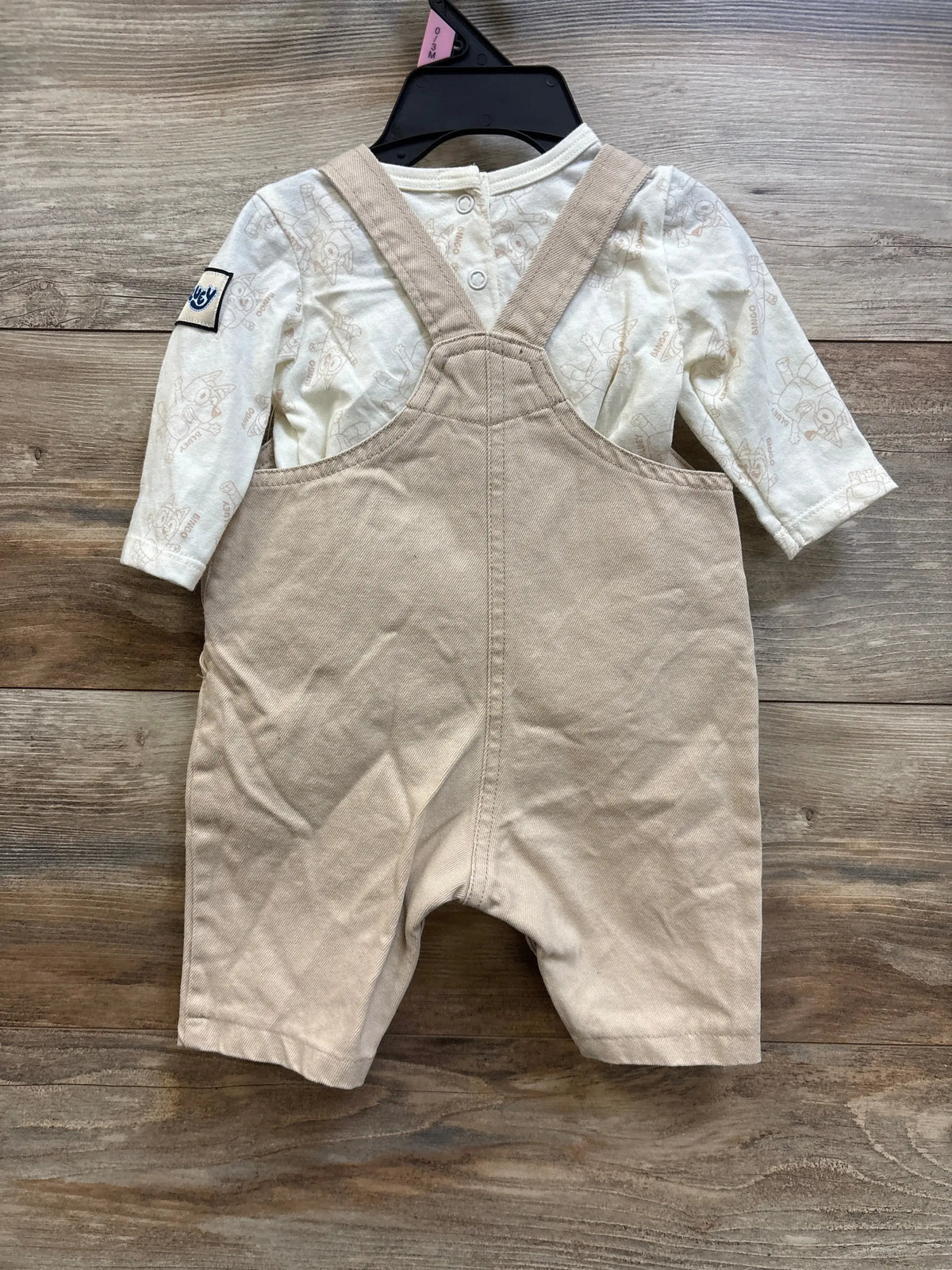 Back view of baby two-piece Bluey set showing tan embroidered overalls over an ivory long-sleeve shirt on a wooden background