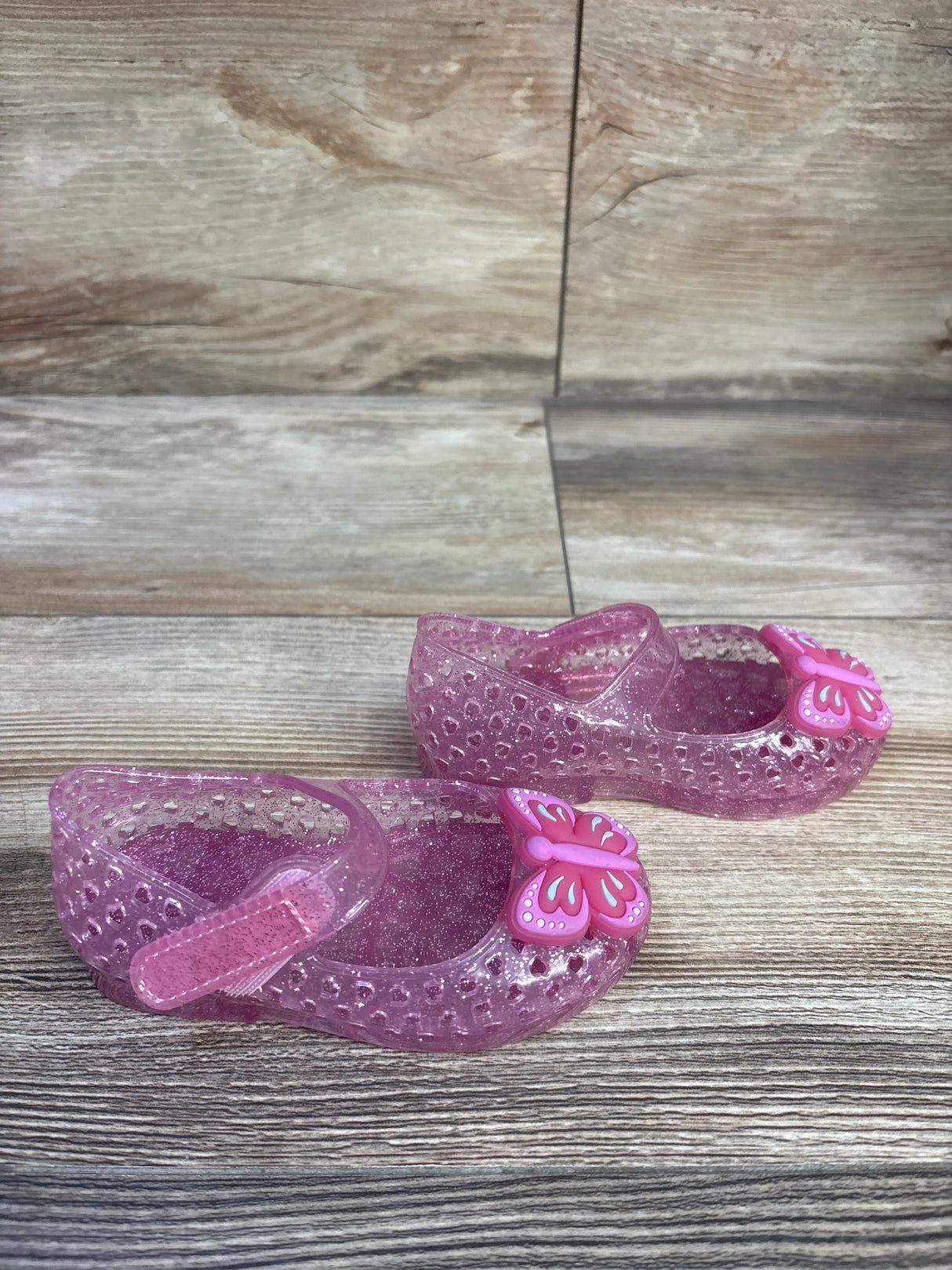 Three-quarter view of pink glitter jelly Mary Jane flats with butterfly charm and hook-and-loop strap on wood floor