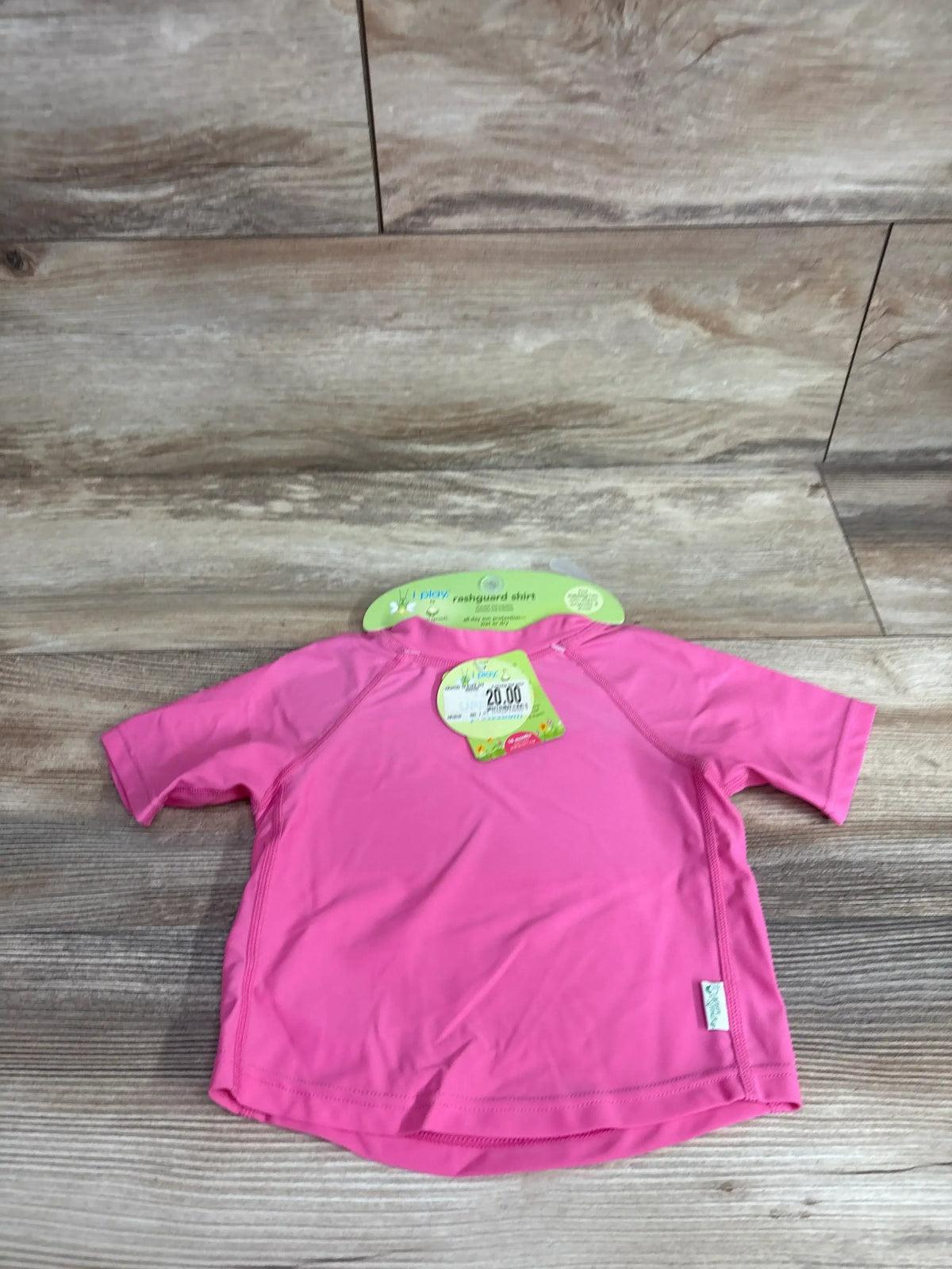 Flat lay of short-sleeve rashguard shirt showcasing bright hot pink color, Green Sprouts hang tag, and size 18m on wood plank background