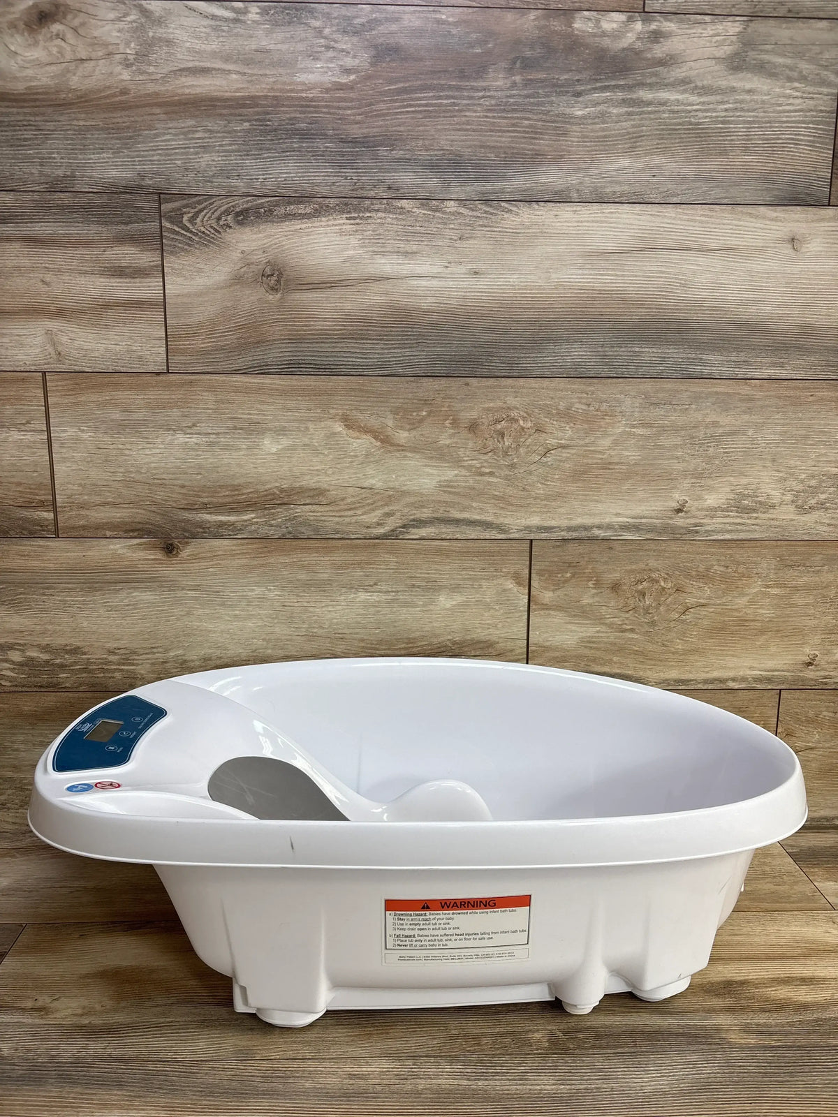 White plastic baby bathtub with built-in scale and warning label on wood panel floor and wall