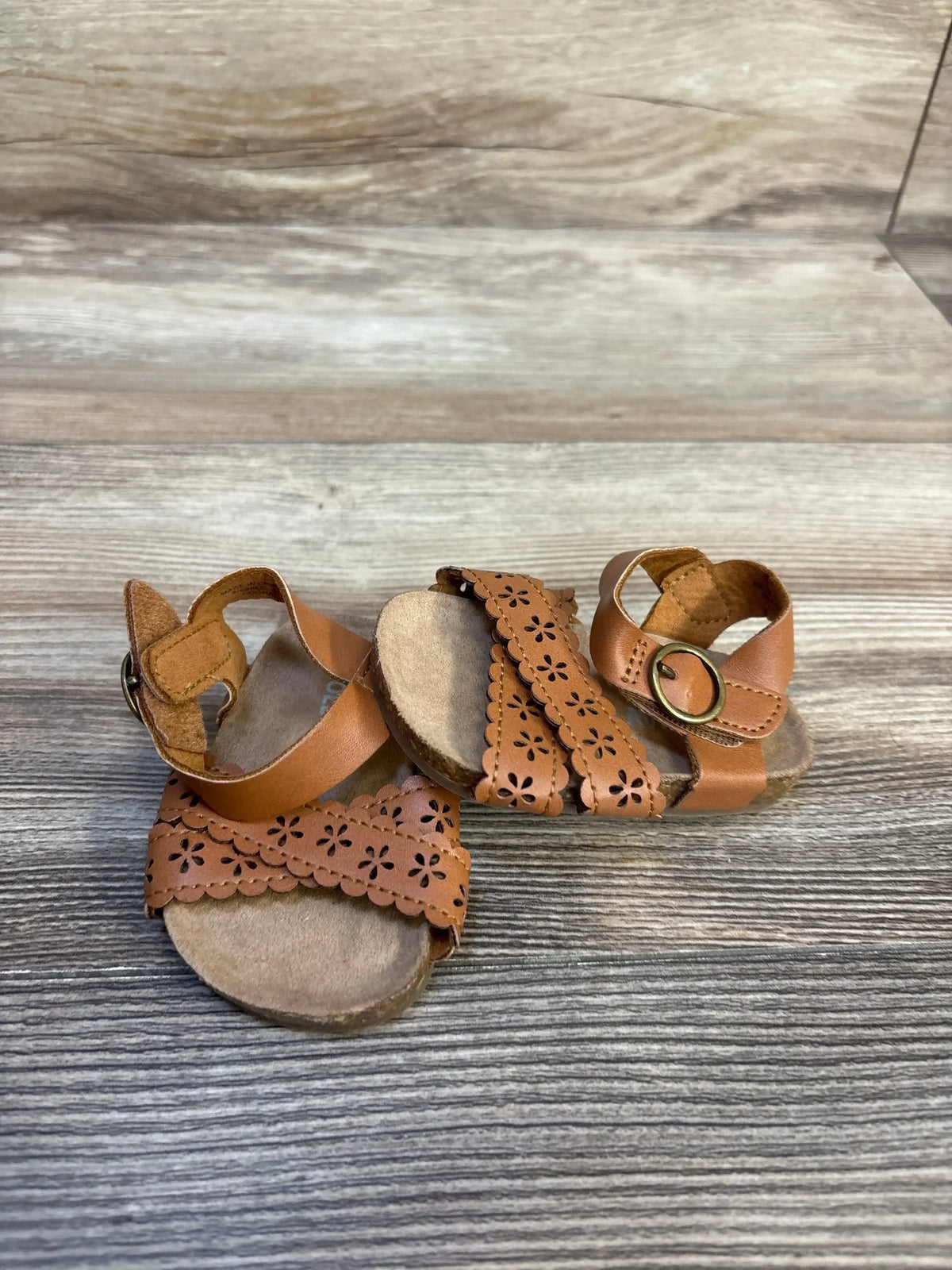 Front view of Old Navy perforated faux-leather baby sandals, beige size 1/2C, floral cutout straps and ankle buckle on wood