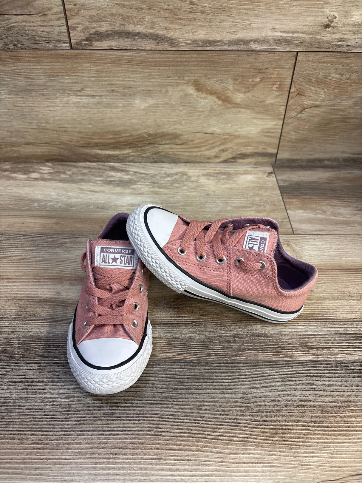 Three-quarter view of pink Converse Chuck Taylor All Star low-top toddler sneakers size 12C showing white rubber toe caps, black foxing stripe, and metal eyelets on a wood plank background