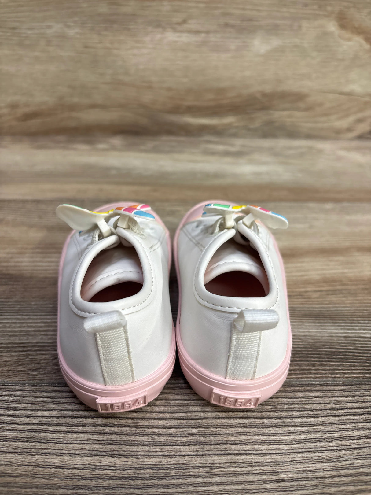 Back view of white kids shoes with pink soles and rainbow decorations on wooden floor