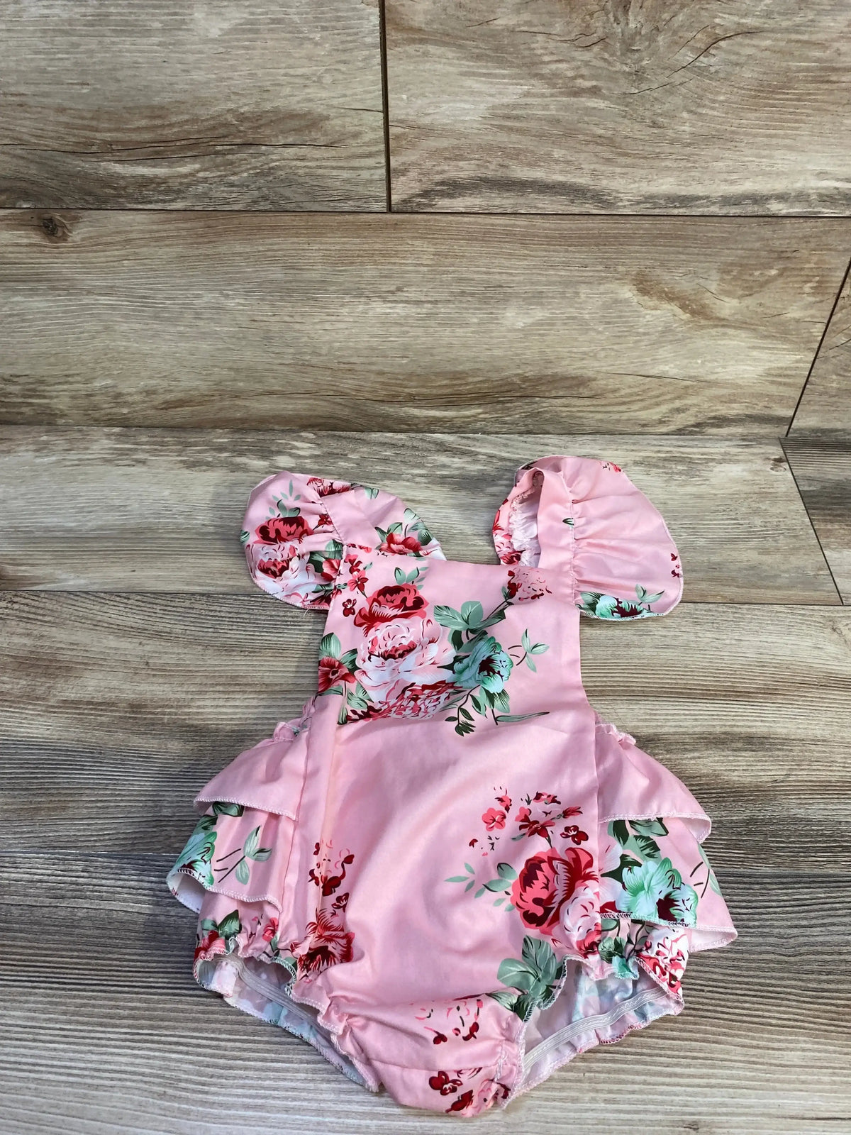 Flat lay of pink floral ruffle bubble romper showing puff sleeves, layered ruffle hips, and elastic leg openings on wooden background