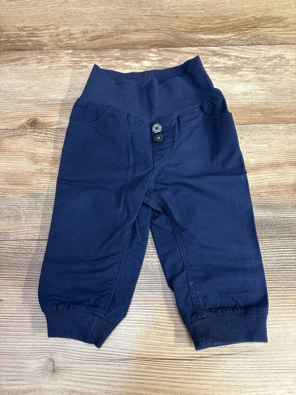 Front view of navy H&M pull-on baby jogger pants size 6–9 months showing elastic ribbed waistband, faux button detail and ribbed cuffs on wood background