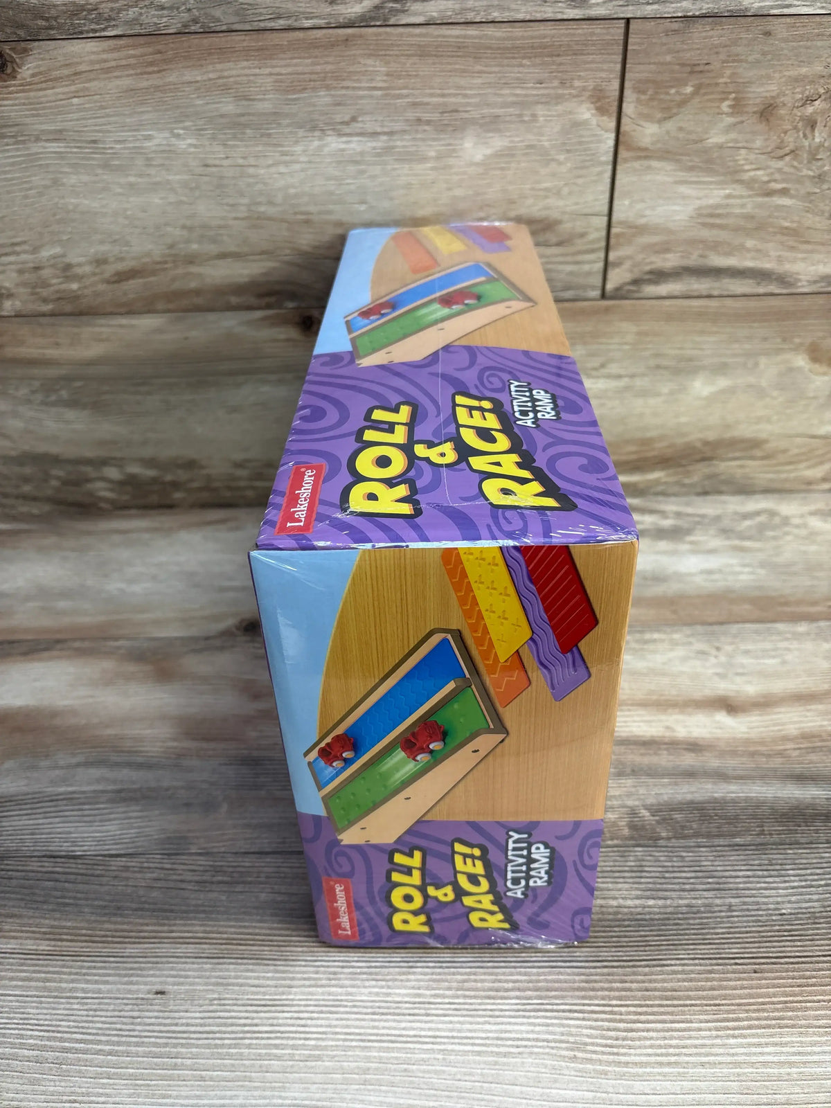 Angled top-front view of boxed Lakeshore Roll & Race activity ramp showing purple packaging and illustration on wood surface
