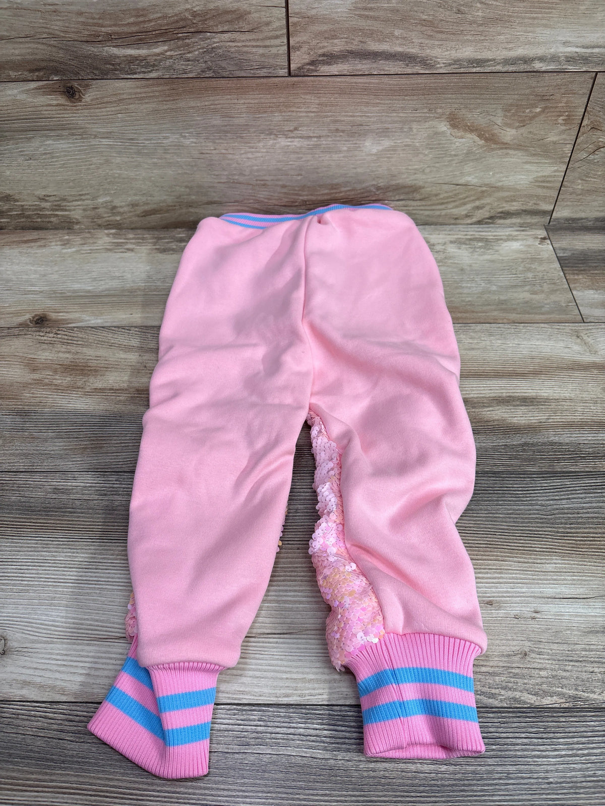 Flat lay of Lola + The Boys Pretty In Pink Sequin Joggers with sequin side panels and striped cuffs on wood-look flooring