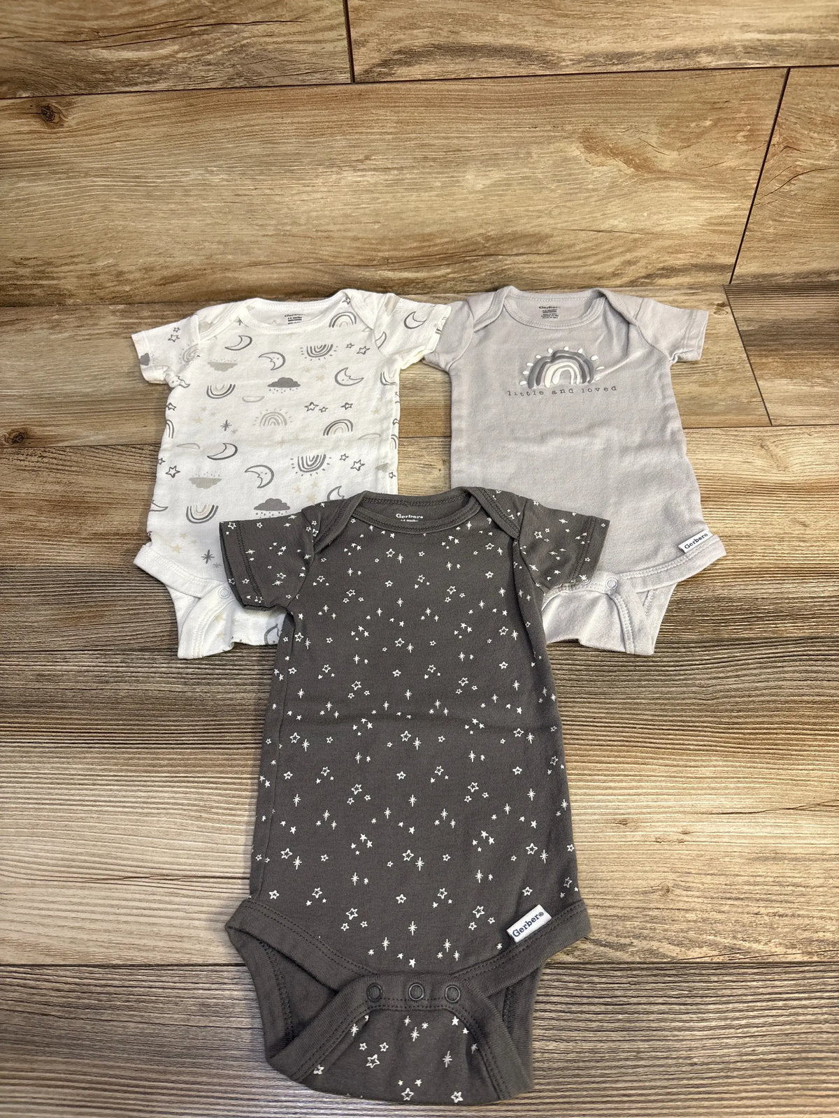 Top-down view of Gerber 3-pack grey bodysuits with moon, rainbow, and star prints on a wood floor