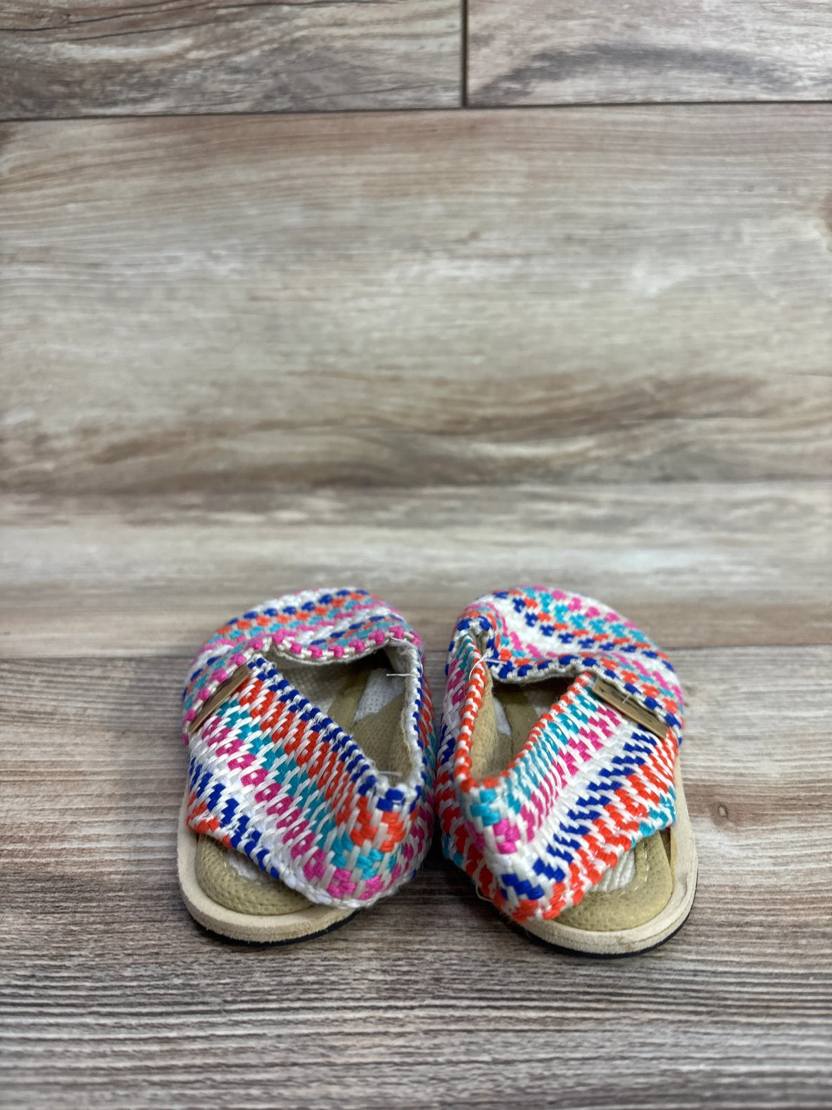Colorful woven baby sandals with pink, orange, blue, and white patterns on wooden floor