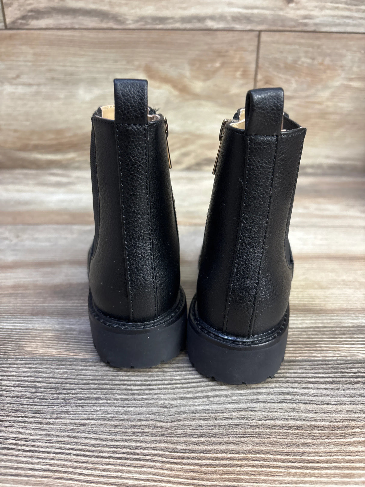 Rear view of Janie & Jack Chelsea boot in black showing back pull tabs, stacked heel, and chunky rubber sole on wooden floor