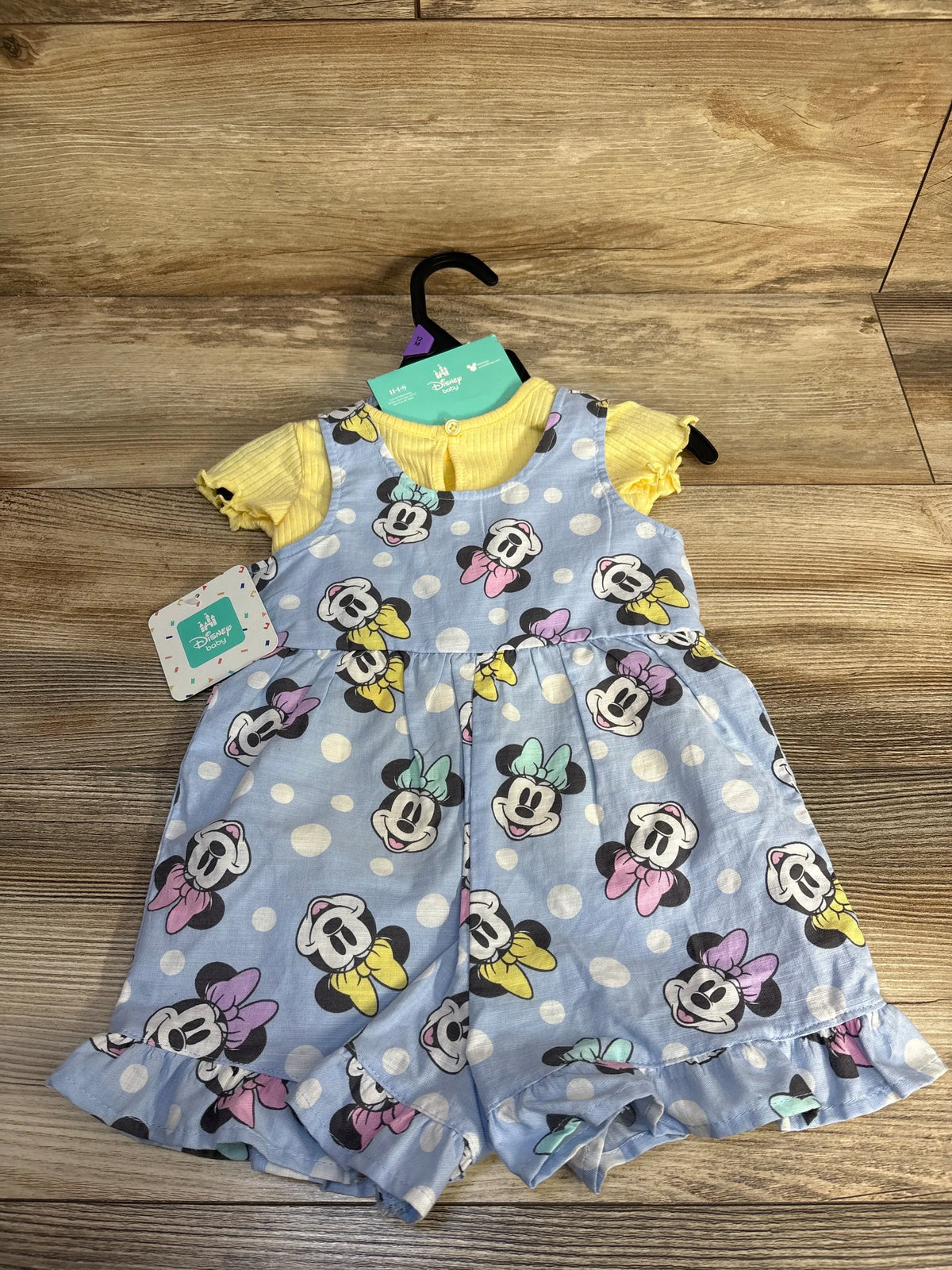 Front view of Disney Baby Minnie Mouse shortall set with yellow ribbed shirt, blue Minnie print, and ruffled hems on wood floor