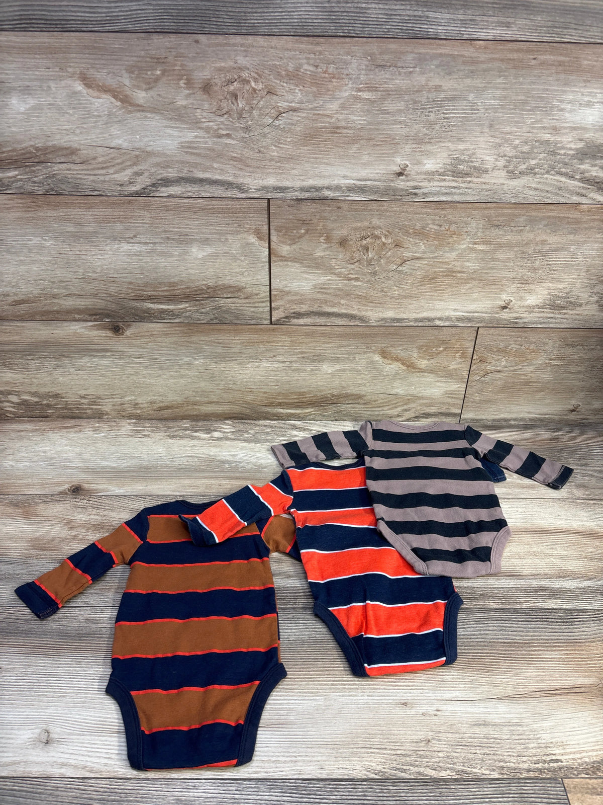 Flat lay of 3 striped long-sleeve baby bodysuits showing navy, orange-red, brown, and gray stripes on a light wood background