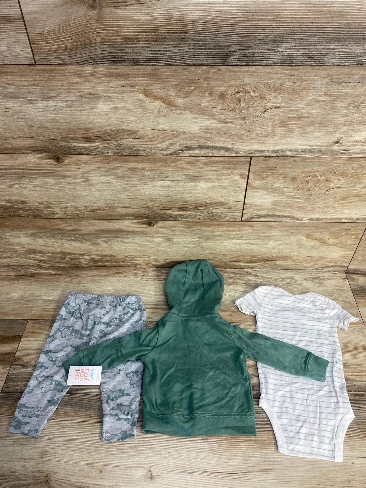Three-piece baby clothing set including gray dinosaur pants, green hooded jacket, and white striped onesie on wooden floor