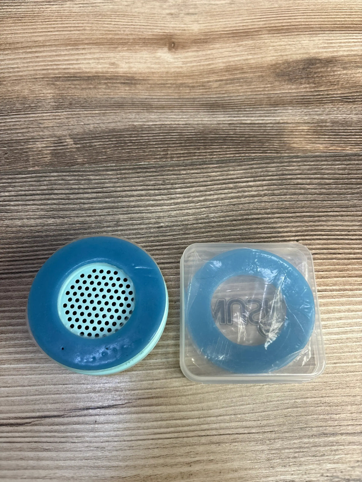 Top view of Wusic WombMusic Bluetooth belly speaker in blue showing perforated speaker grill and matching silicone belly ring in plastic case on wood surface