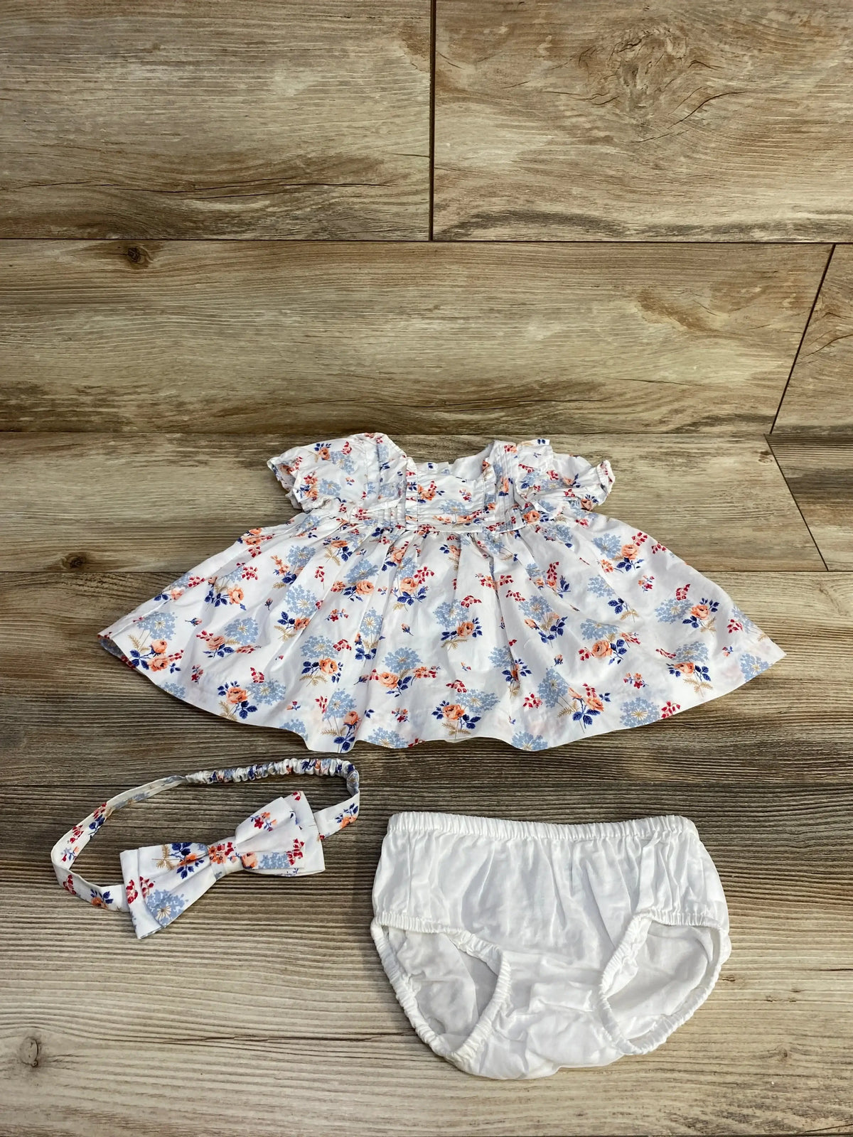 Overhead view of white floral pintuck baby dress set showcasing pintuck bodice and ruffled sleeves with matching headband and white bloomers on wood background