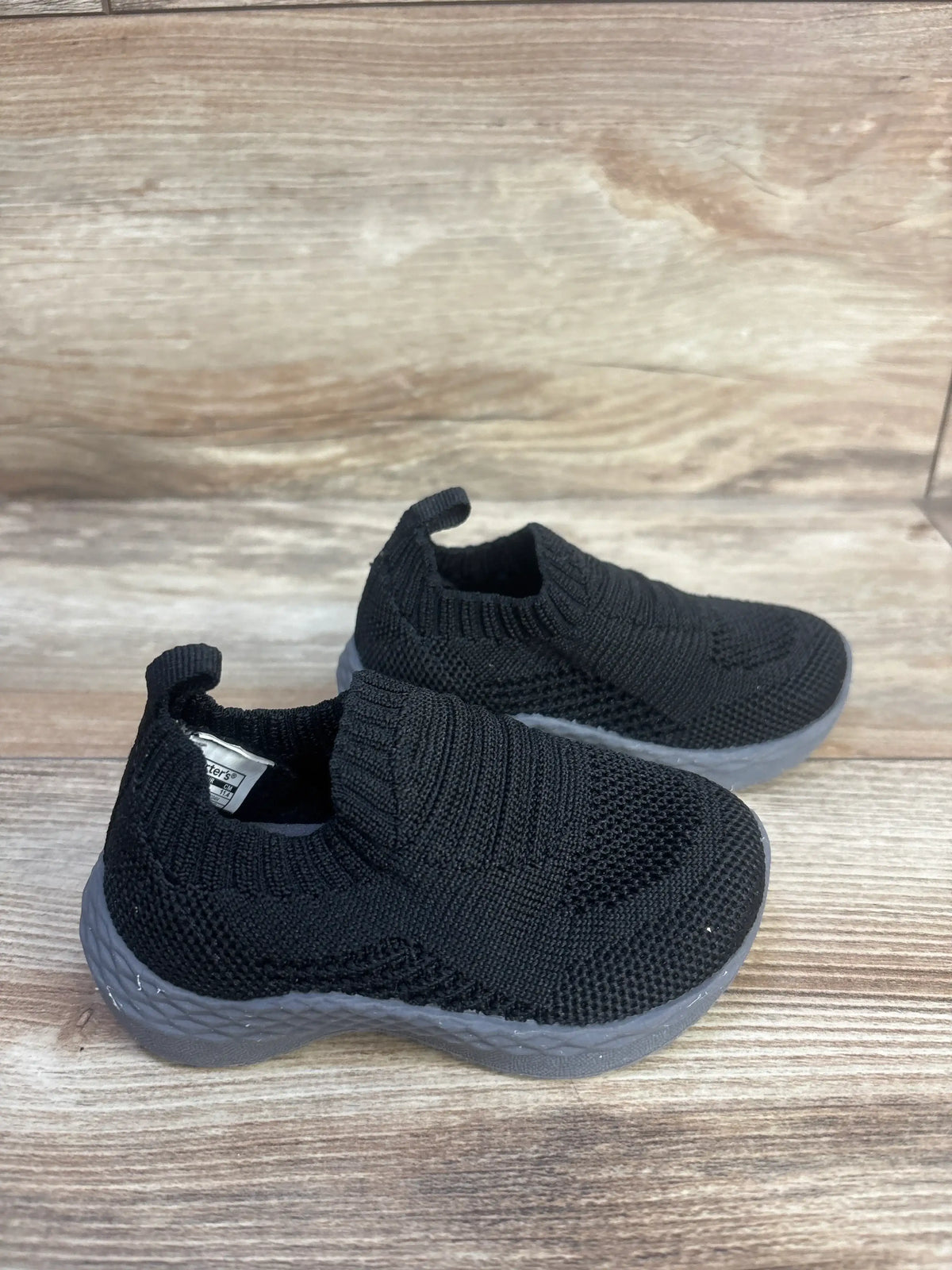 Angled view of Carter’s Taylen pull-on athletic sneakers showcasing black knit uppers and gray grippy soles on a wood surface