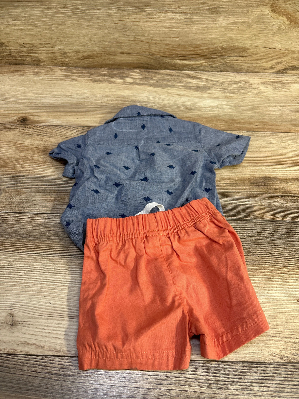 Flat lay of 2-piece button-up shirt and elastic-waist shorts, blue printed top and orange shorts, size 3m on wood background