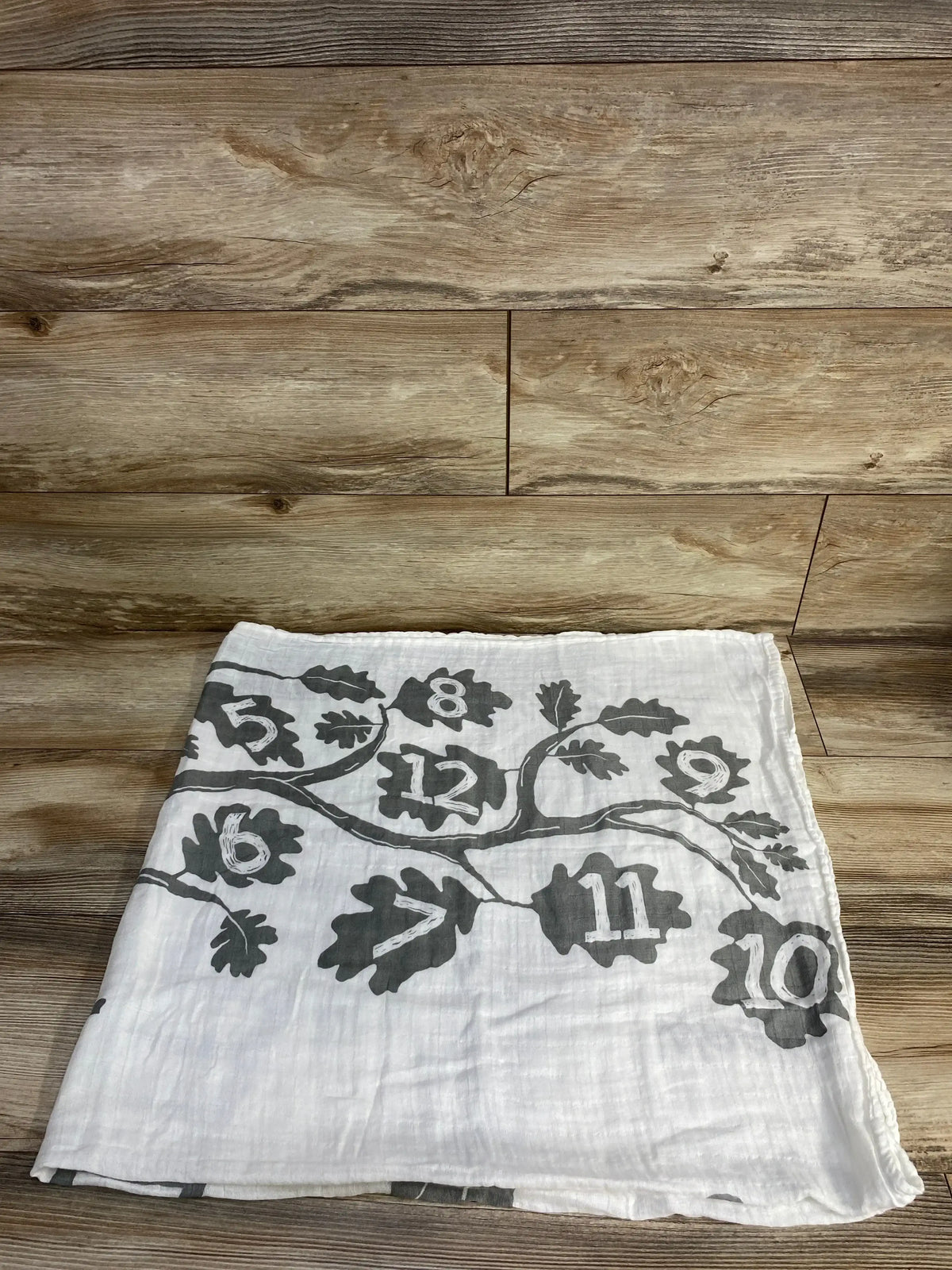 Folded front view of Baby Wings Height Milestone Blanket, Tree showcasing gray tree and leaf print with numbers 5 to 12 on wooden floor