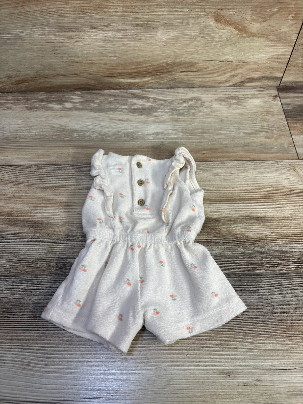 Front view of an ivory floral terry cloth romper showcasing ruffled shoulders and button front with wood-look floor background