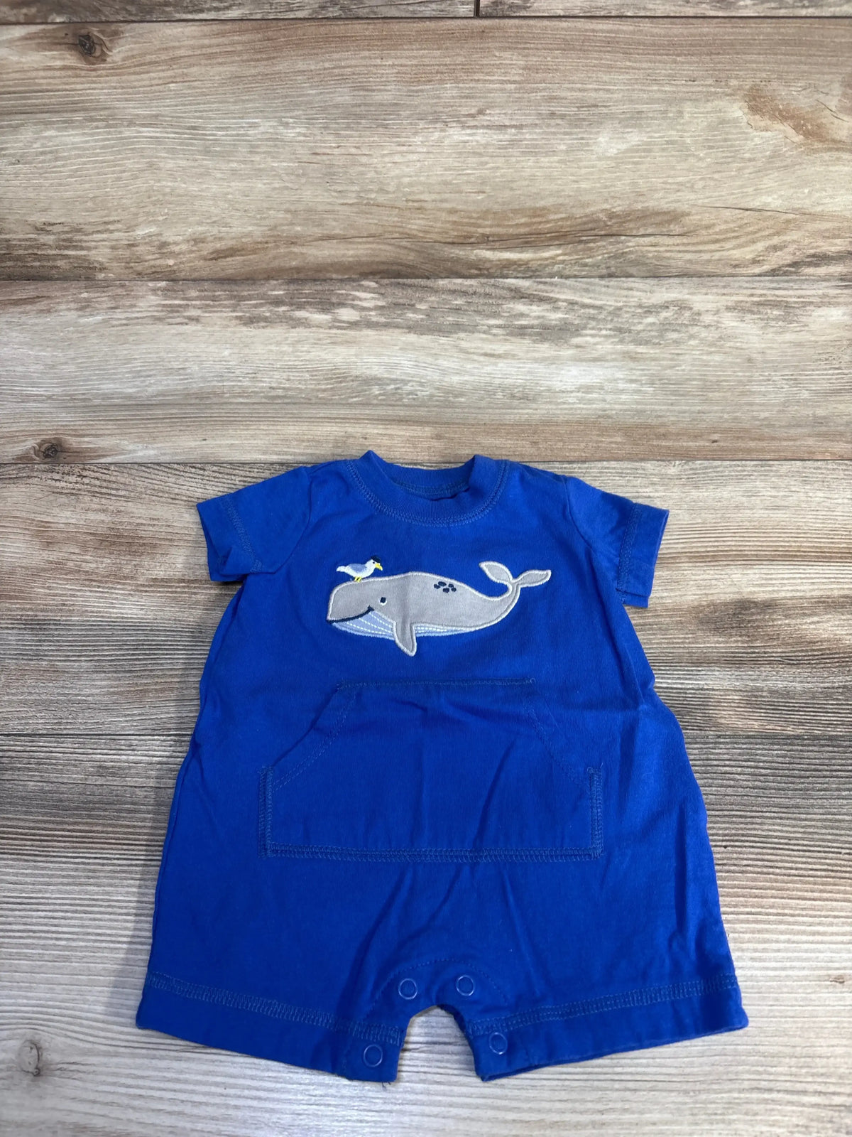 Flat-lay of blue Carter’s shortie romper (3 months) showing grey whale applique and front kangaroo pocket on wood background
