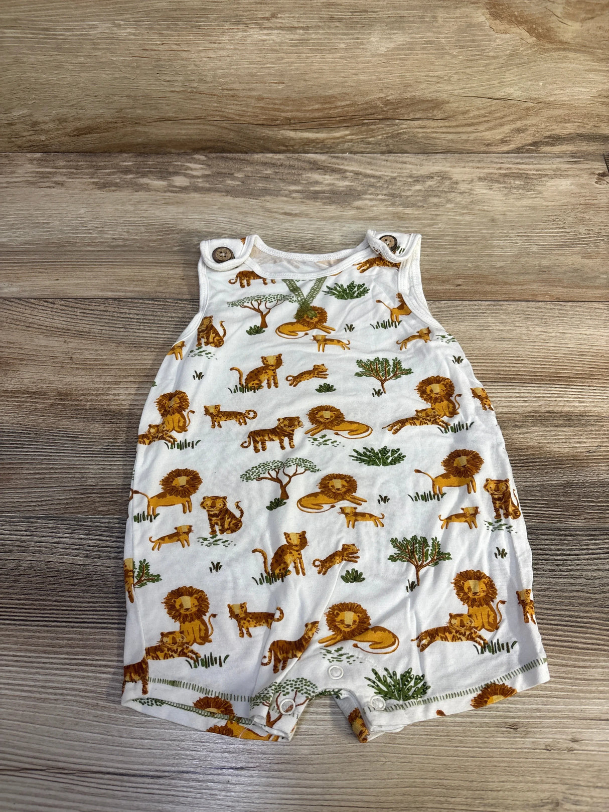 Flat lay of Mudpie white tank shortie romper with lion print, shoulder button snaps and snap crotch on wood floor background
