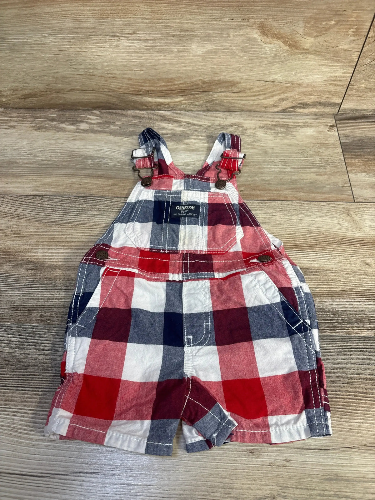 Front flat lay of Oshkosh shortalls 12M in red, navy and white plaid with adjustable metal buckle straps, bib pocket on wood floor