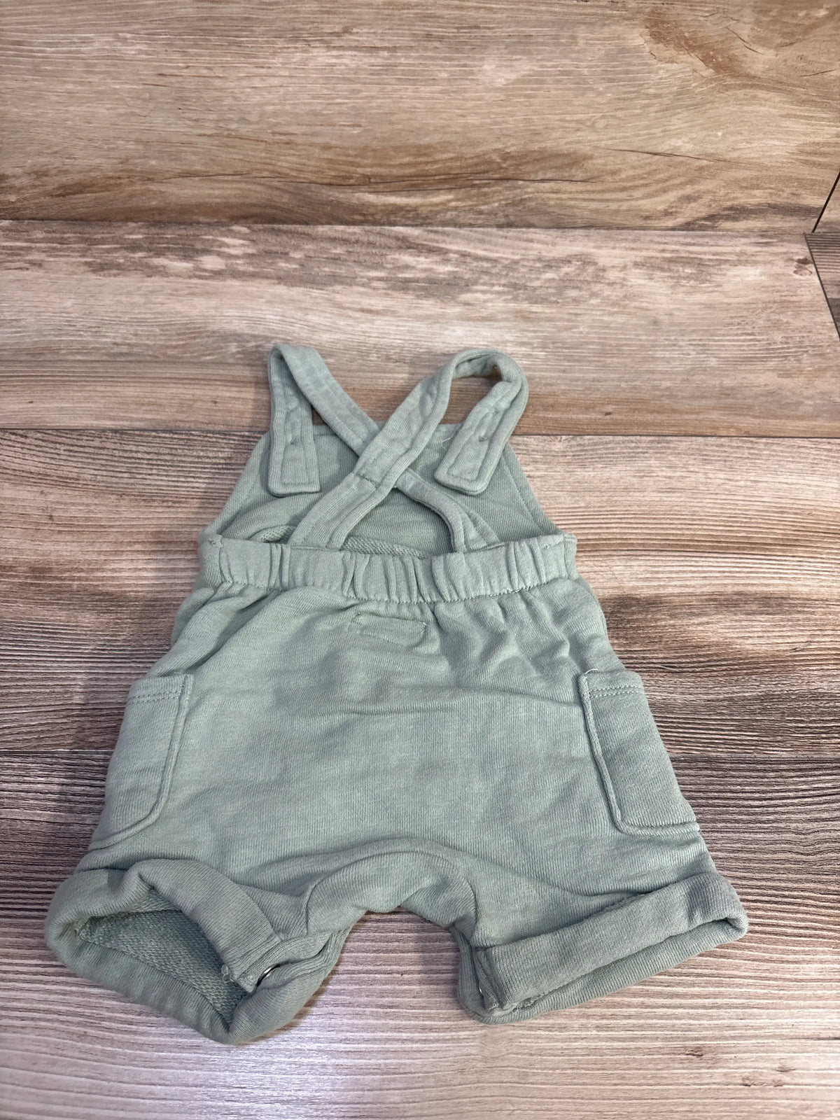 Top down view of green baby shortalls size 3M with crossed straps, elastic waist and front pockets on wood background