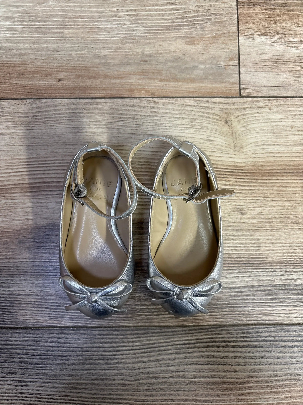 Pair of silver toddler Mary Jane shoes with ankle straps and bows on wooden floor