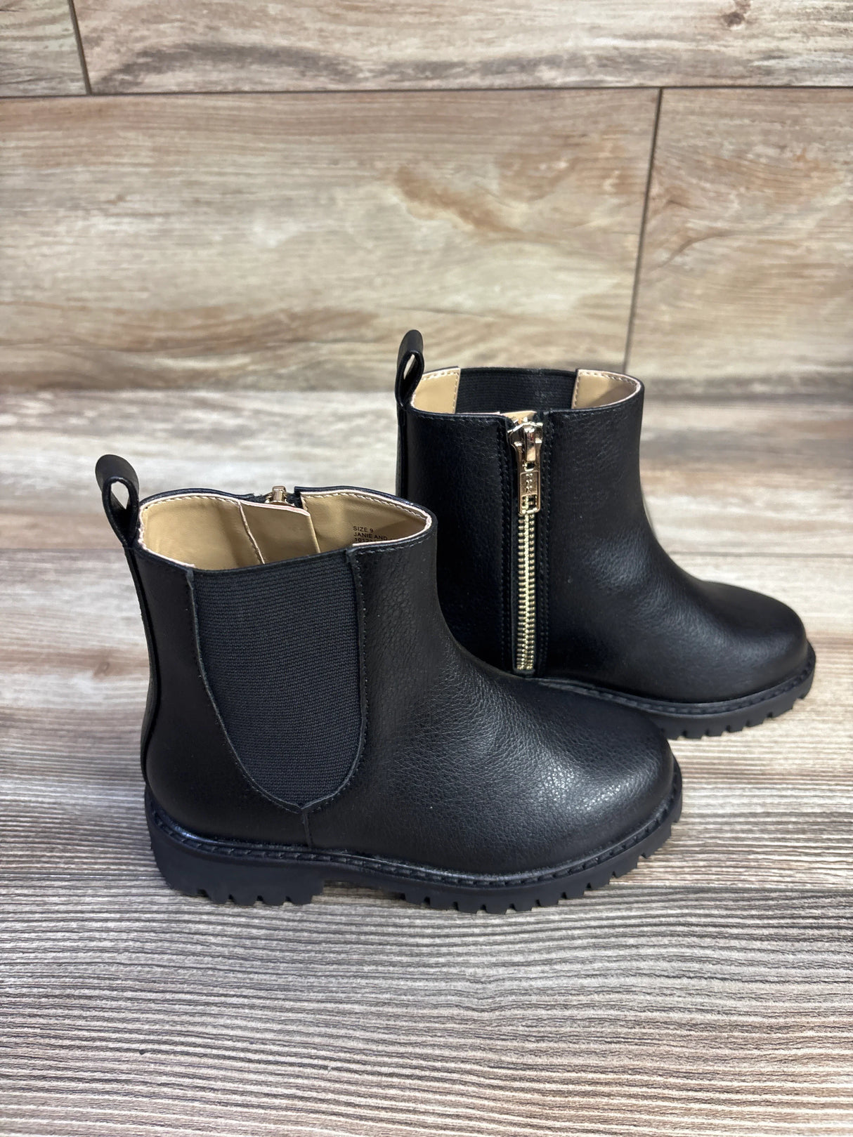 Three quarter view of toddler black Chelsea boots showing elastic side panel, pull tab and gold zipper on wood floor